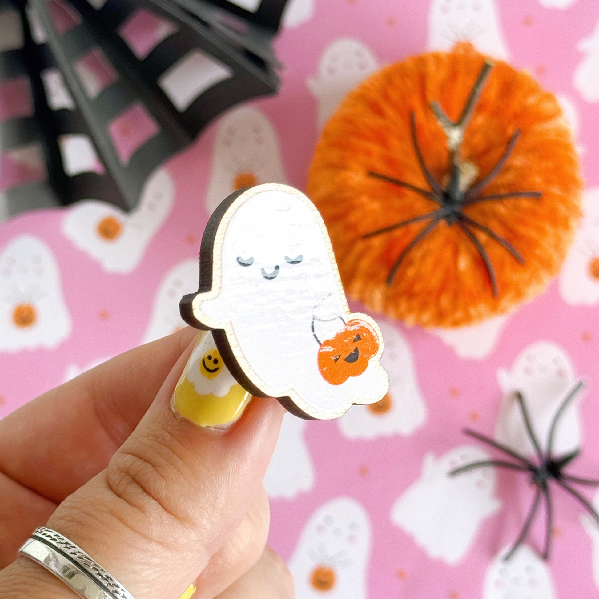 A wooden pin badge, of a cute smiling ghost, holding a trick or treating tub which is a pumpkin.