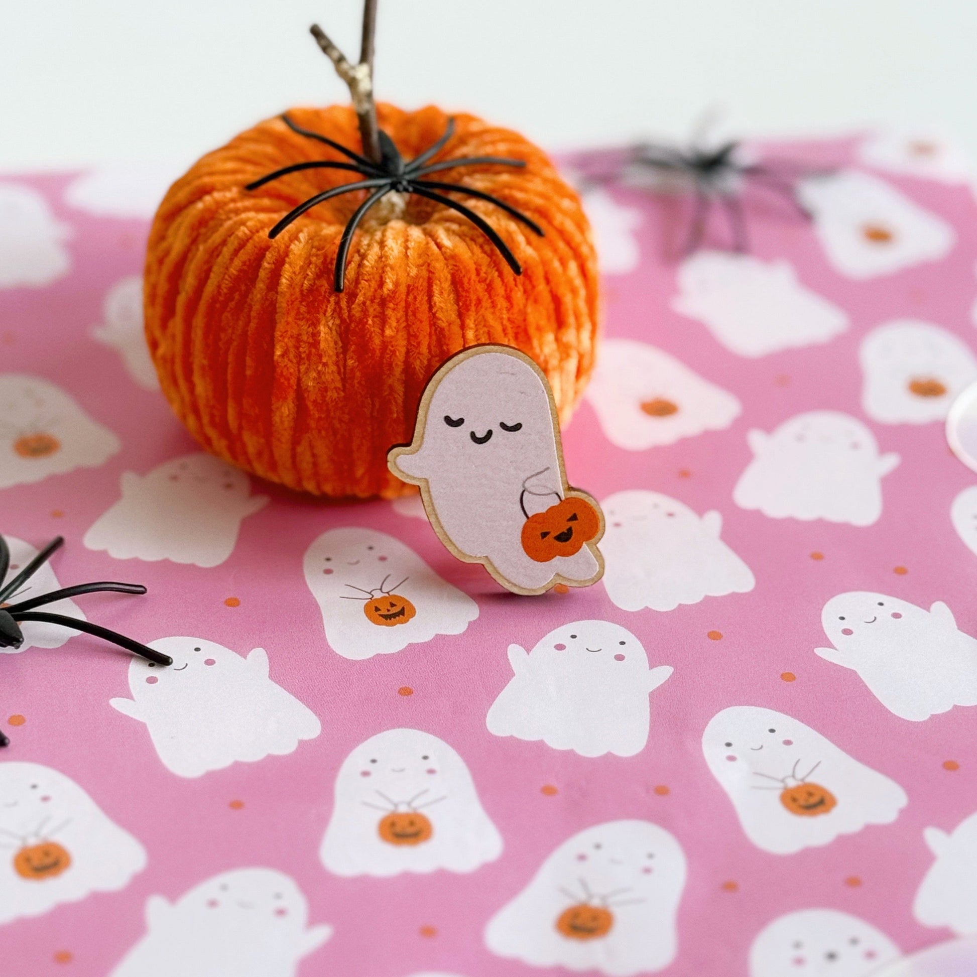 A wooden pin badge, of a cute smiling ghost, holding a trick or treating tub which is a pumpkin.