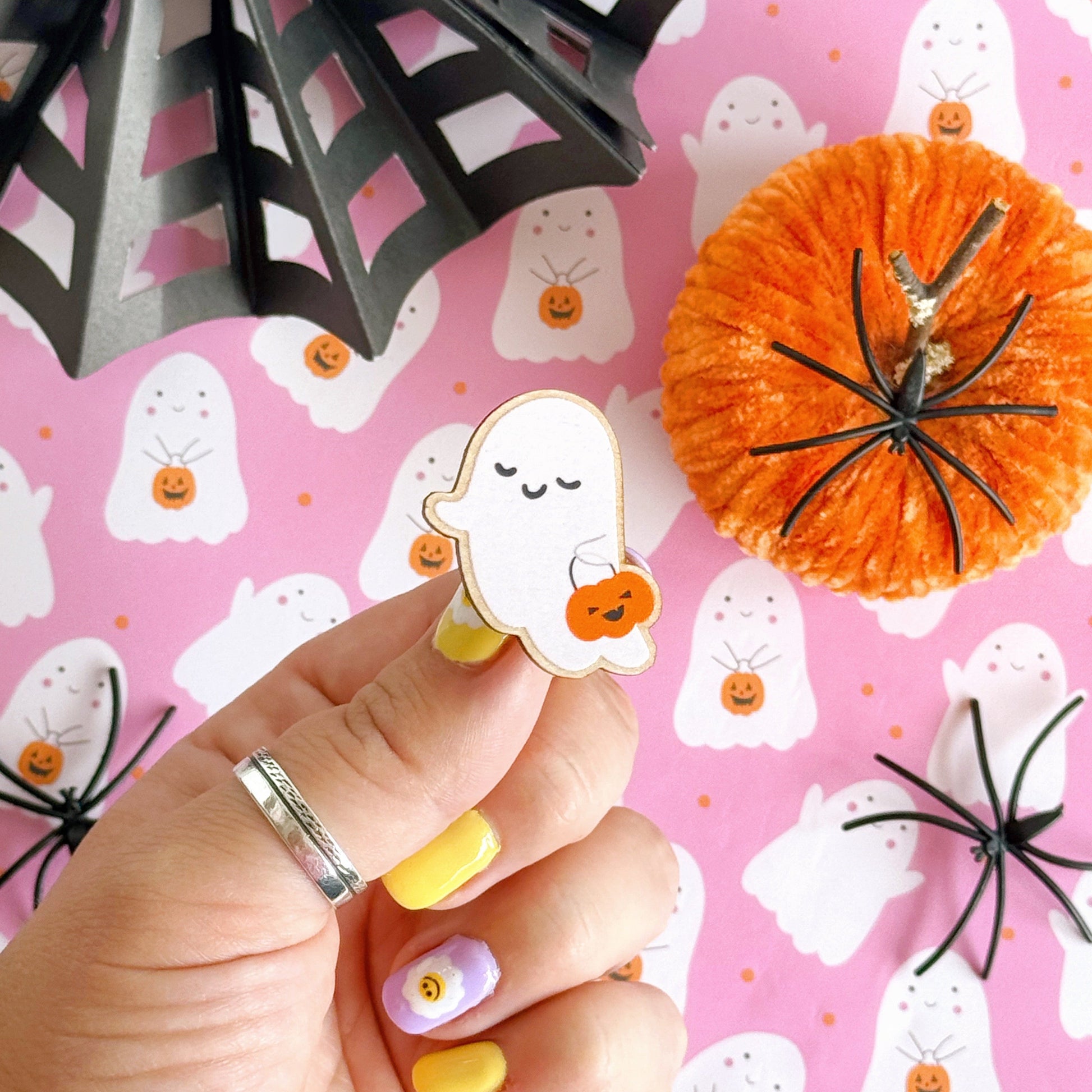 A wooden pin badge, of a cute smiling ghost, holding a trick or treating tub which is a pumpkin.