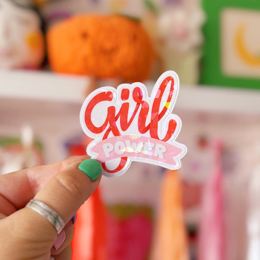 Hand holding a 'Girl Power' sticker in front of a colourful background with toys and decorations.