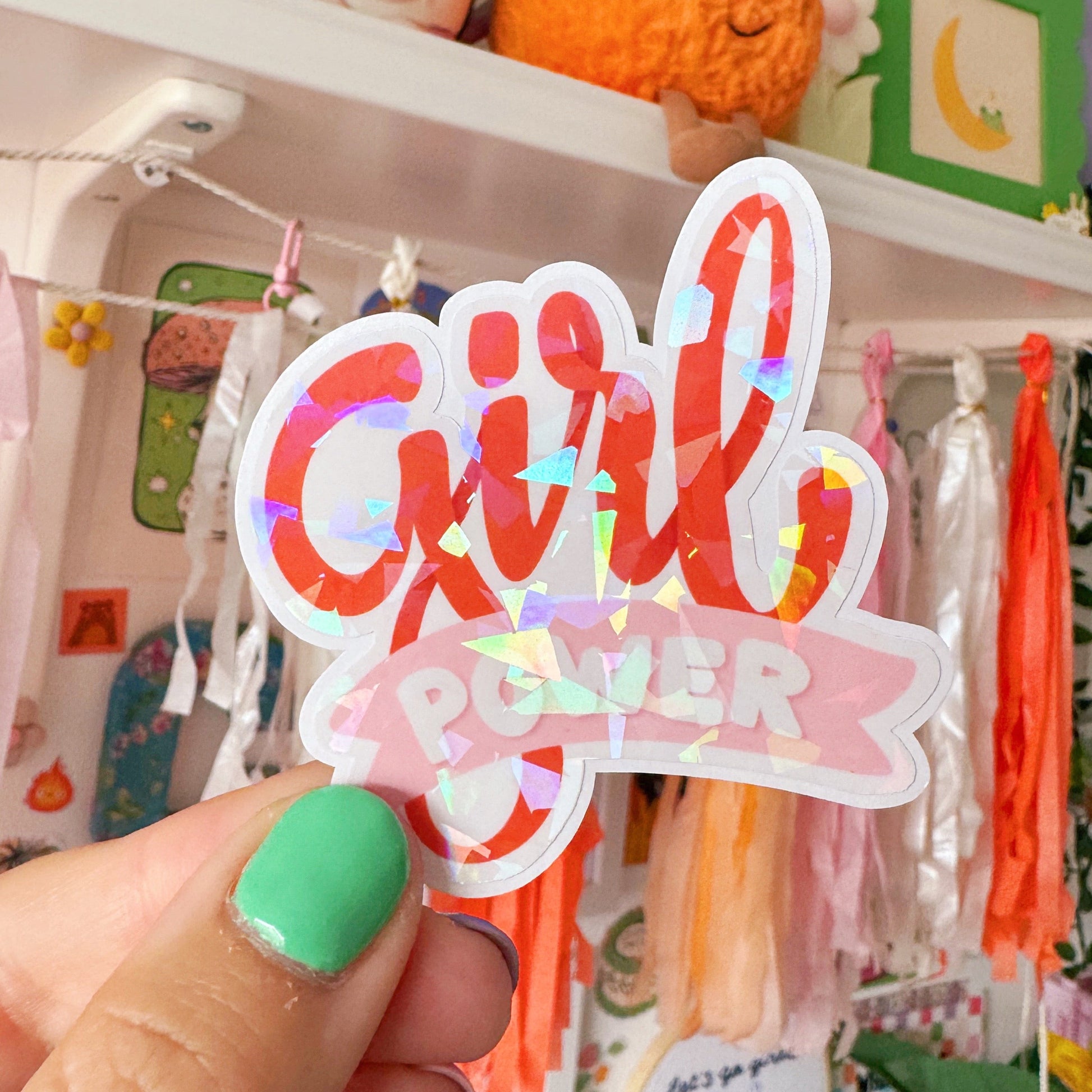 Hand holding a 'Girl Power' sticker in front of a shelf with various items