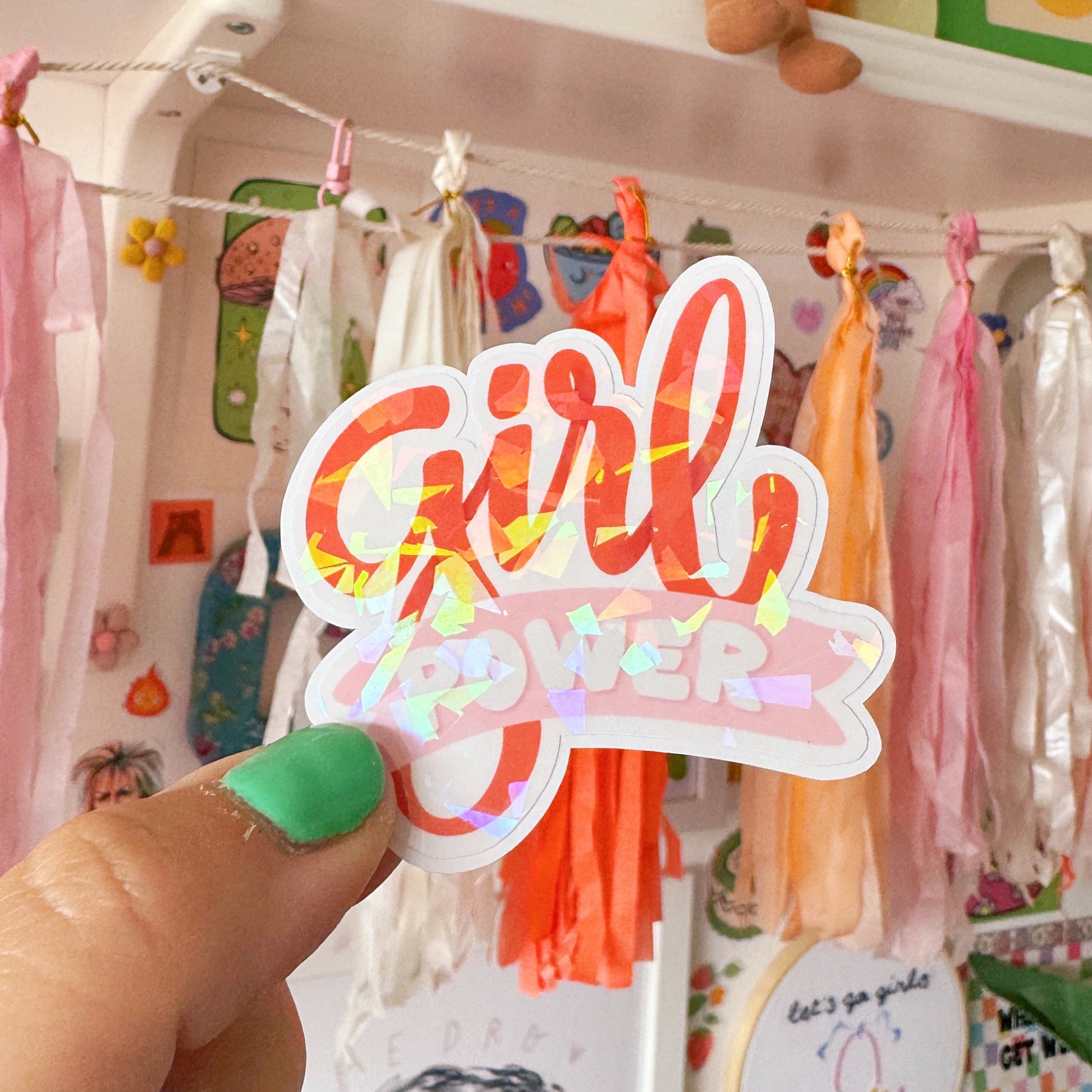 Hand holding a 'Girl Power' sticker in front of a colourful shelf with toys and decorations.