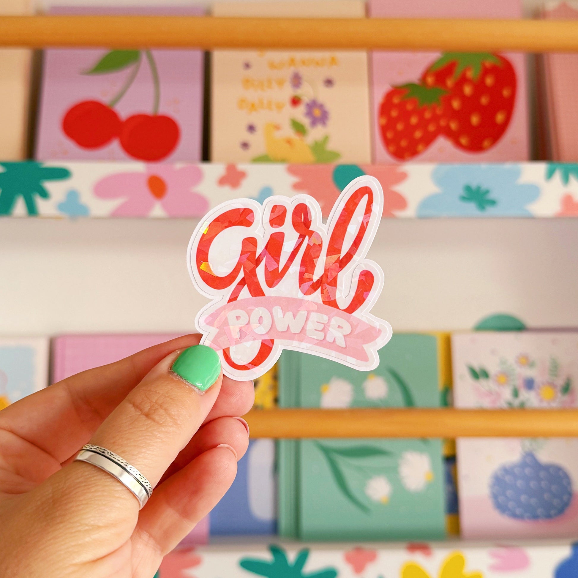 Hand holding a 'Girl Power' sticker in front of a colorful shelf with stationery items.