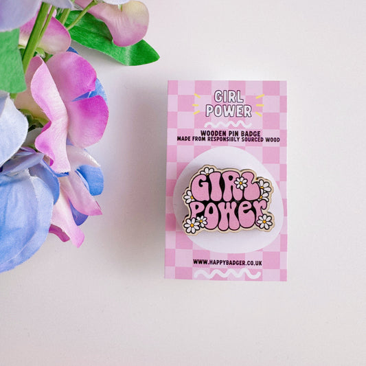 A pink wooden pin badge with the words girl power on, surrounded by white daisies.
