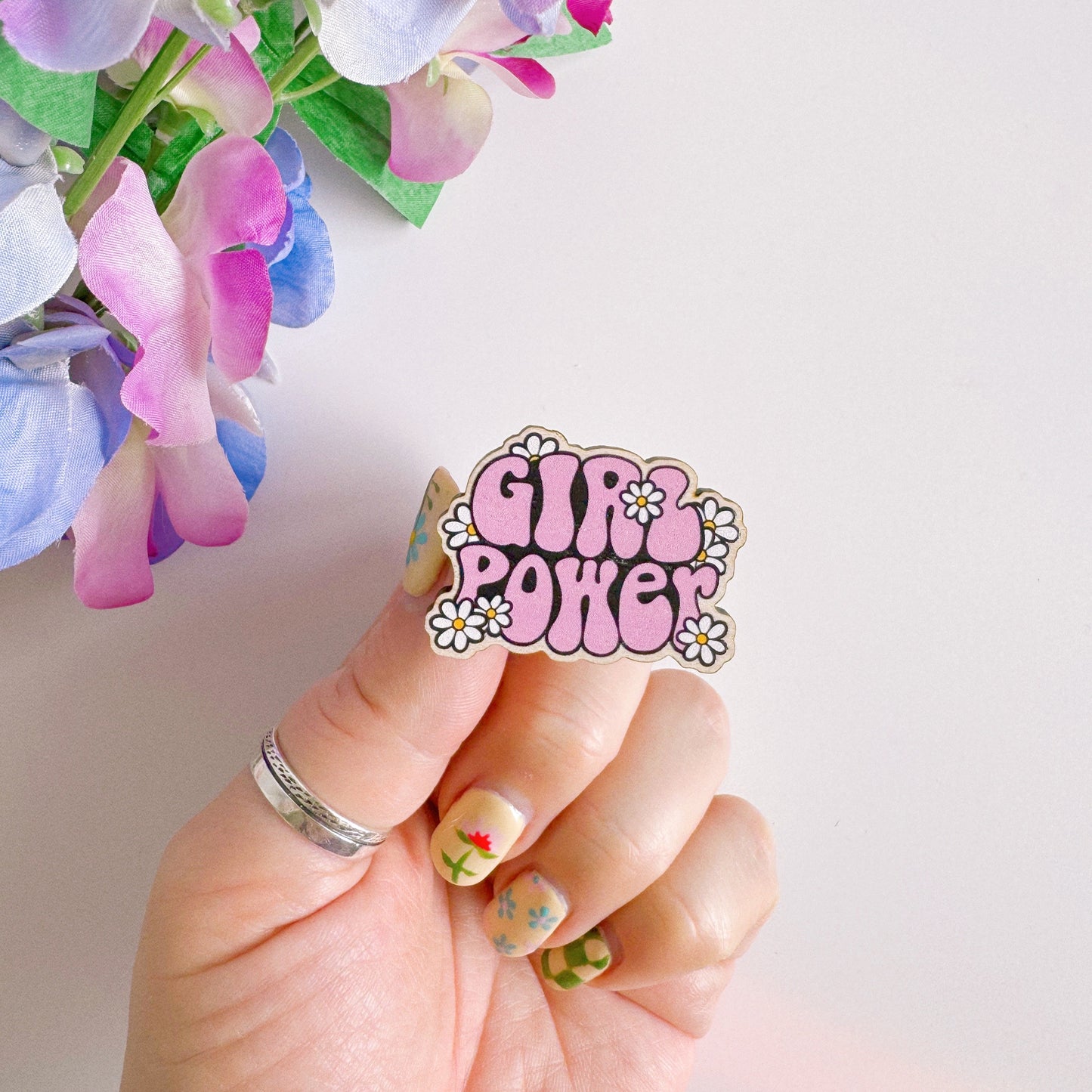 A pink wooden pin badge with the words girl power on, surrounded by white daisies.