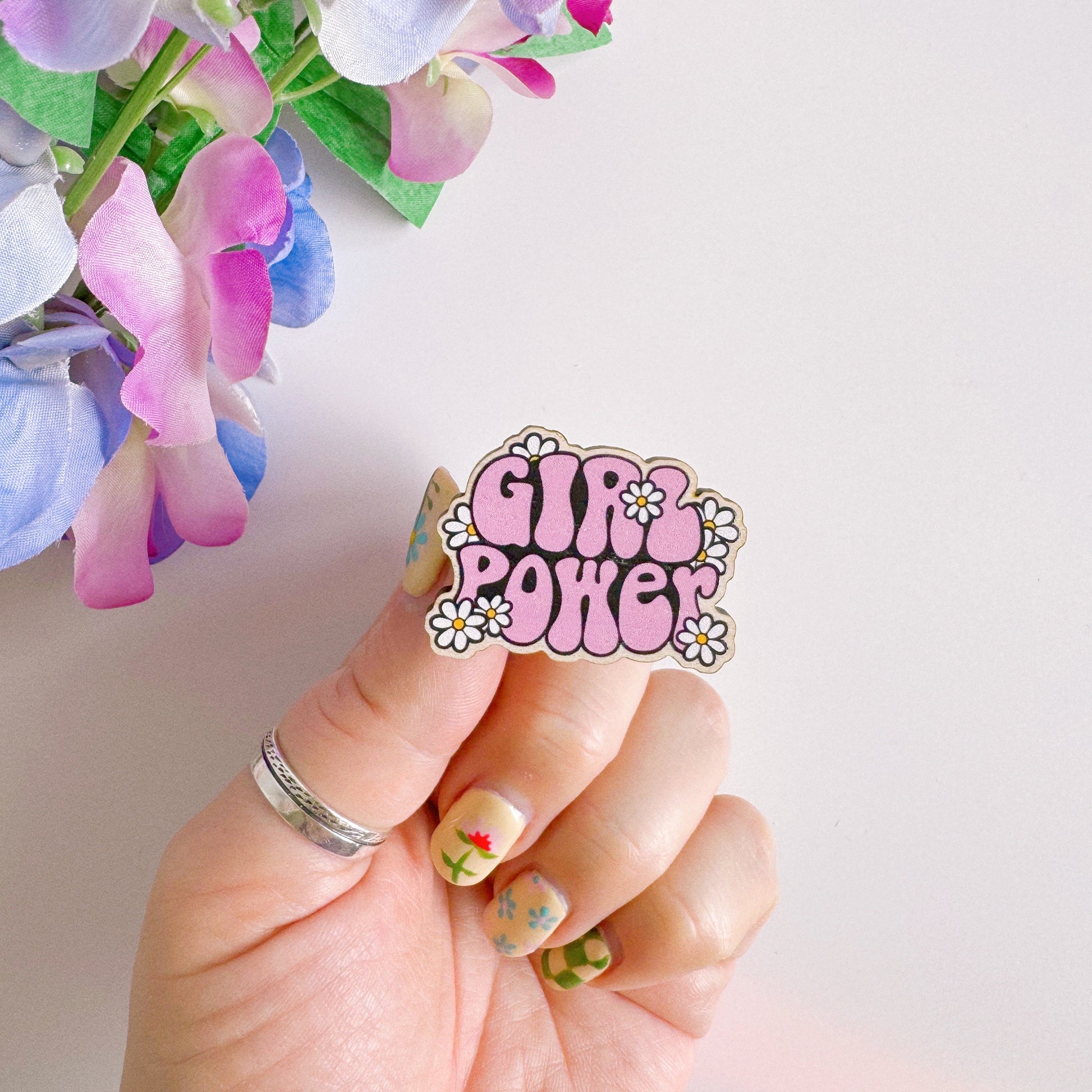 A pink wooden pin badge with the words girl power on, surrounded by white daisies.