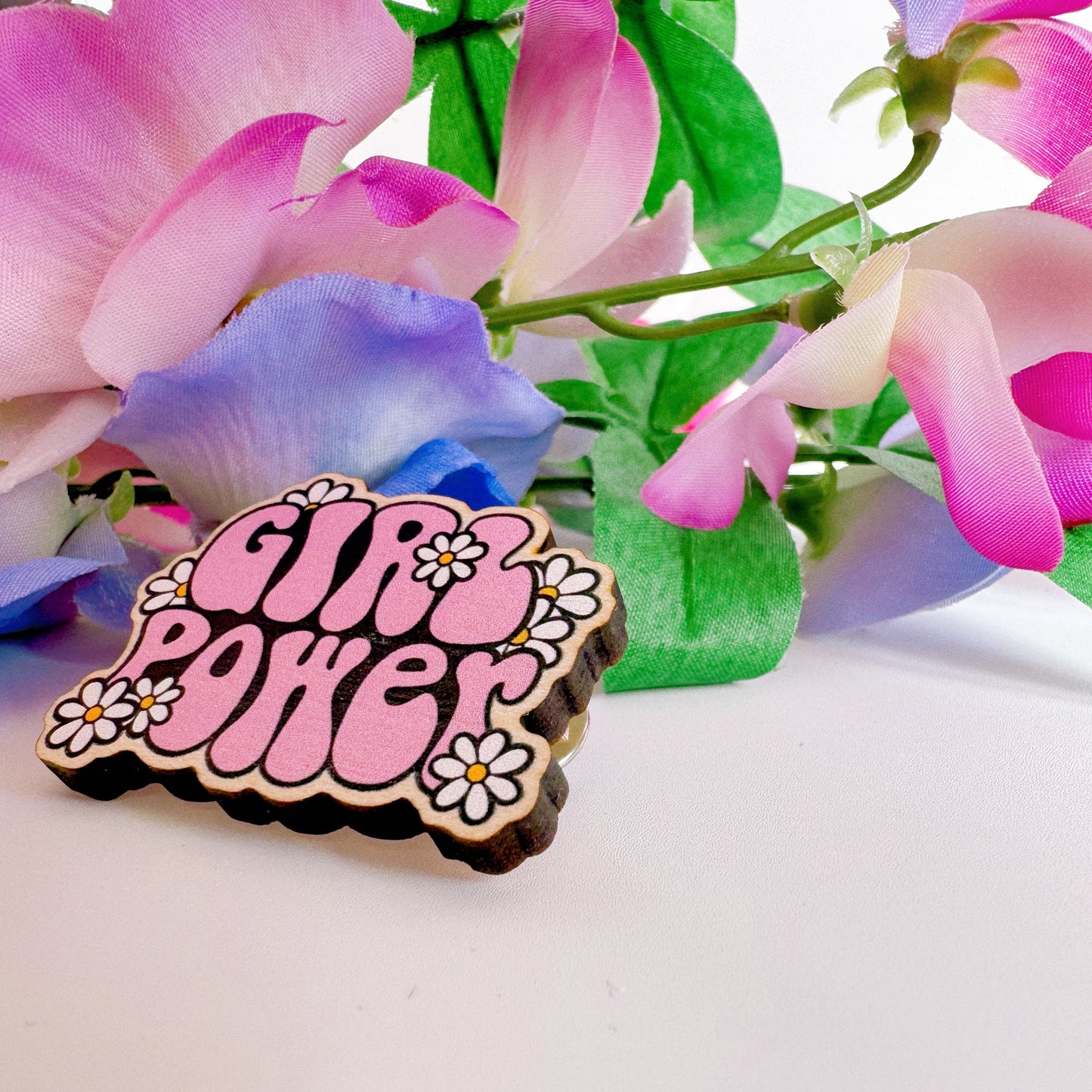 A pink wooden pin badge with the words girl power on, surrounded by white daisies.