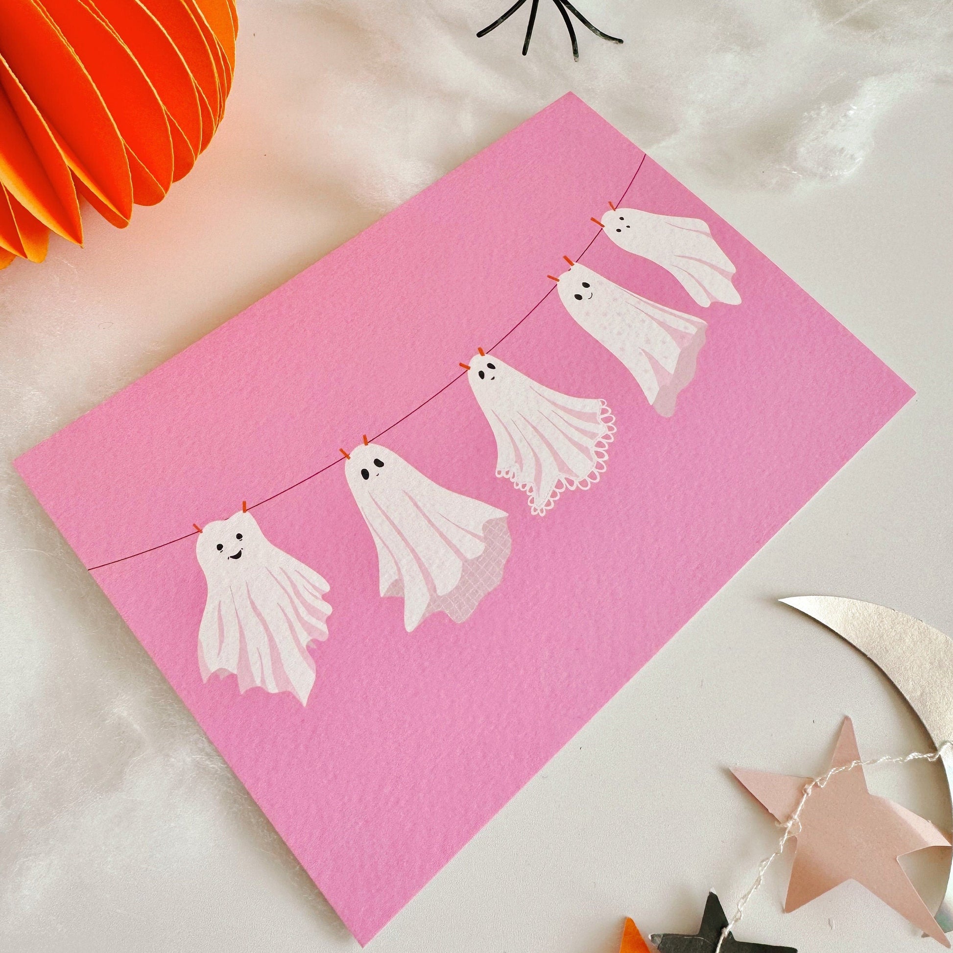 A halloween themed postcard of 5 ghosts hanging on a washing line, on a pink background.