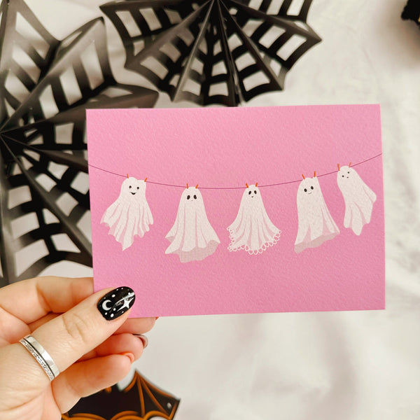 A halloween themed postcard of 5 ghosts hanging on a washing line, on a pink background.