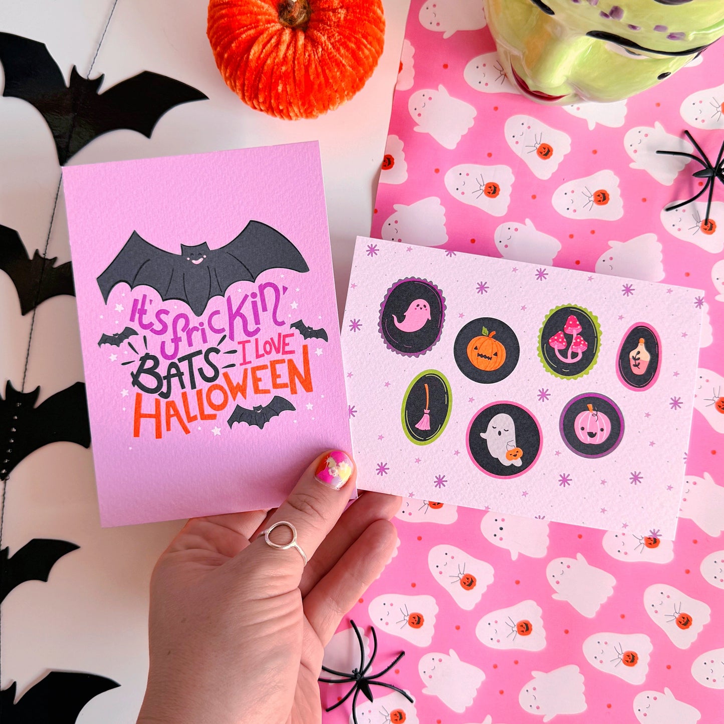 A landscape postcard with 7 illustrated frames, inside of which are spooky cute Halloween illustrations. The background of the postcard is pale purple with purple starbursts.