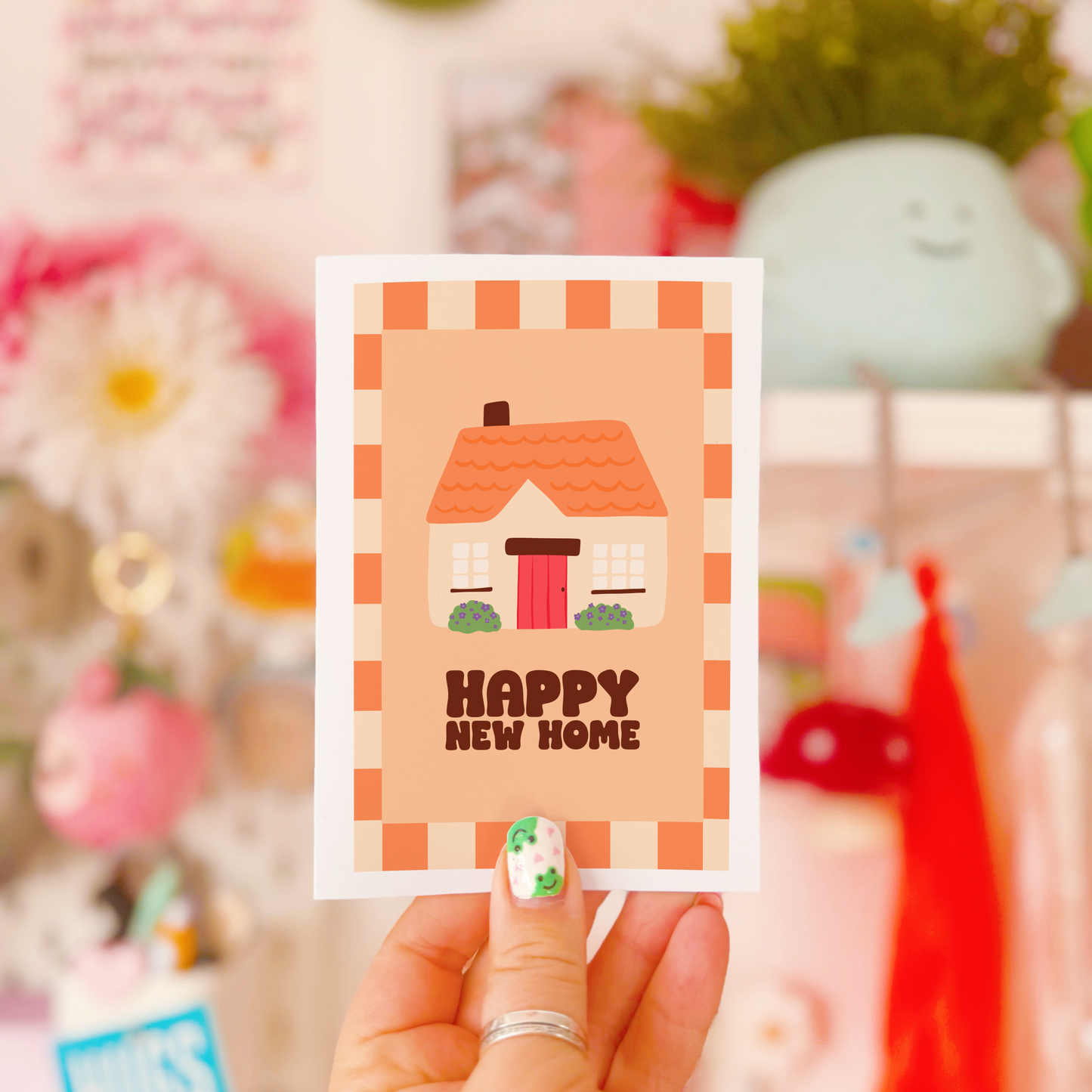 An image of a greetings card, with the message 'Happy New Home' on the front. The card has an orange and cream border, with a cute cosy cottage illustration.