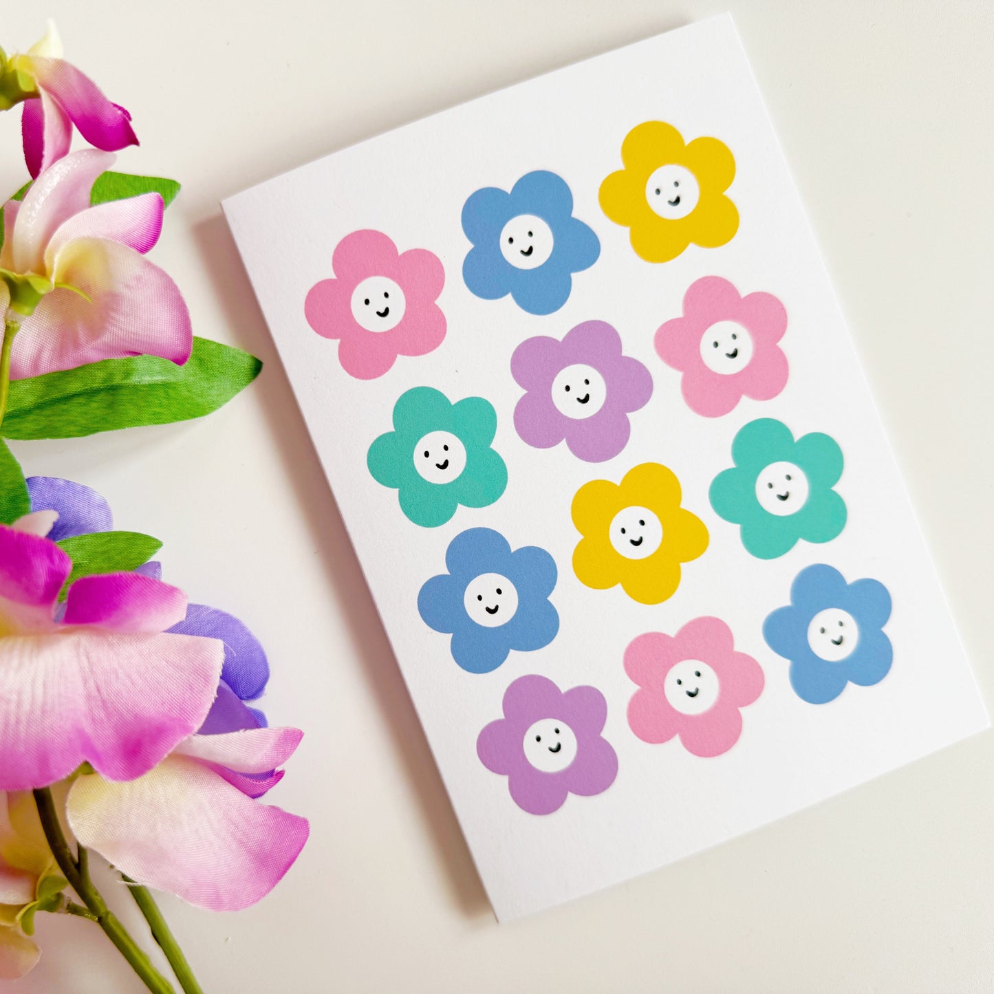 A white card with 12 colourful smiling daisy-like flowers on the front.