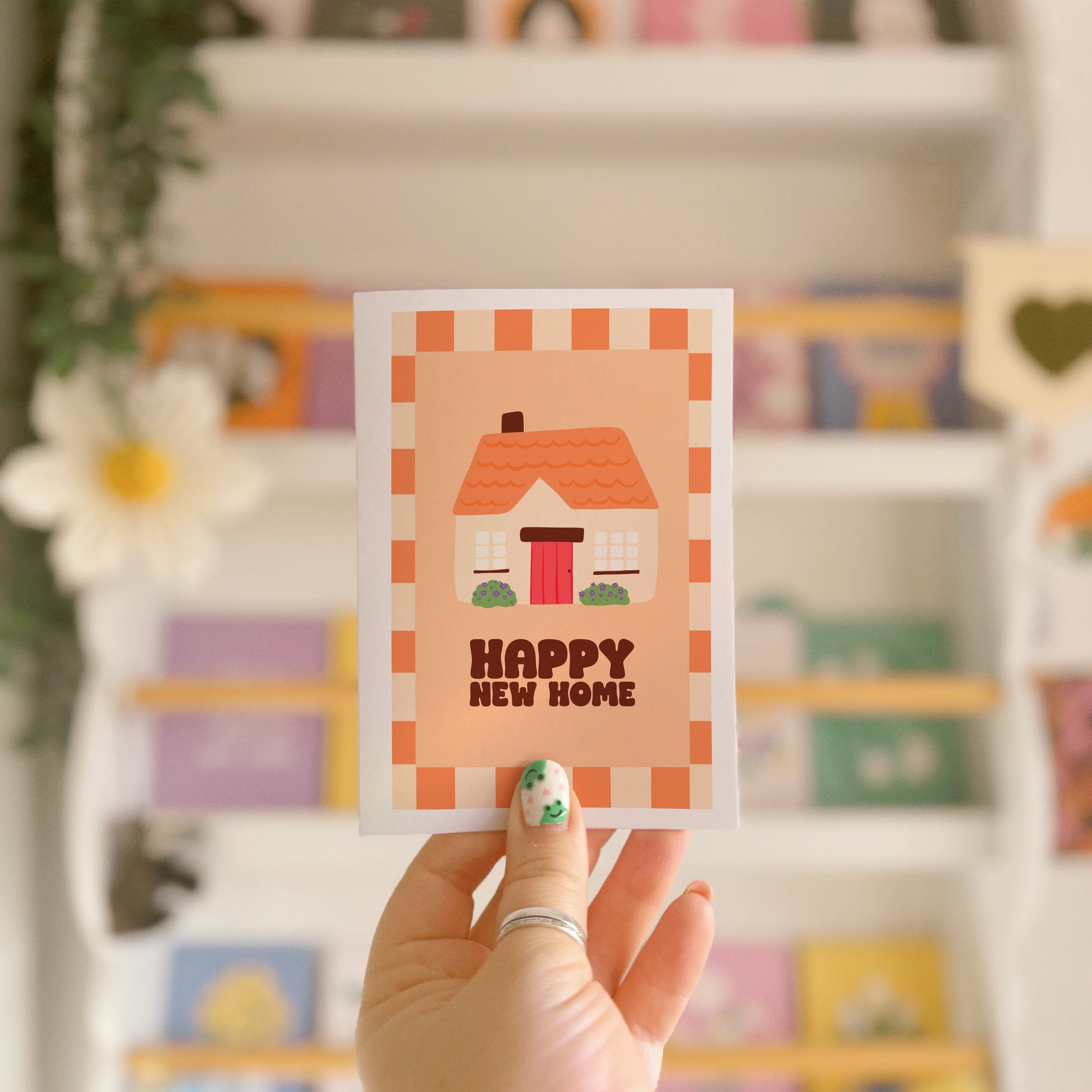 An image of a greetings card, with the message 'Happy New Home' on the front. The card has an orange and cream border, with a cute cosy cottage illustration.