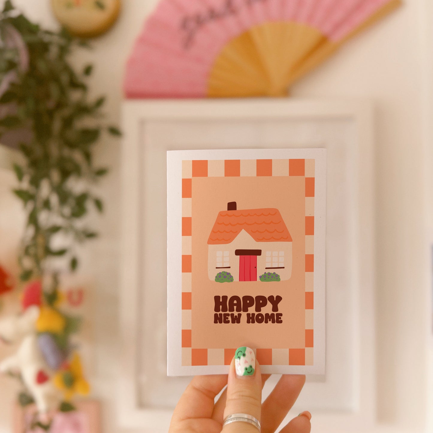 An image of a greetings card, with the message 'Happy New Home' on the front. The card has an orange and cream border, with a cute cosy cottage illustration.