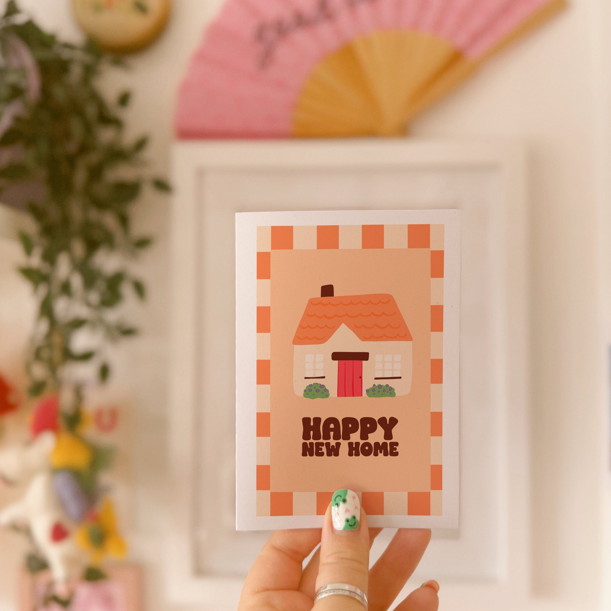 An image of a greetings card, with the message 'Happy New Home' on the front. The card has an orange and cream border, with a cute cosy cottage illustration.