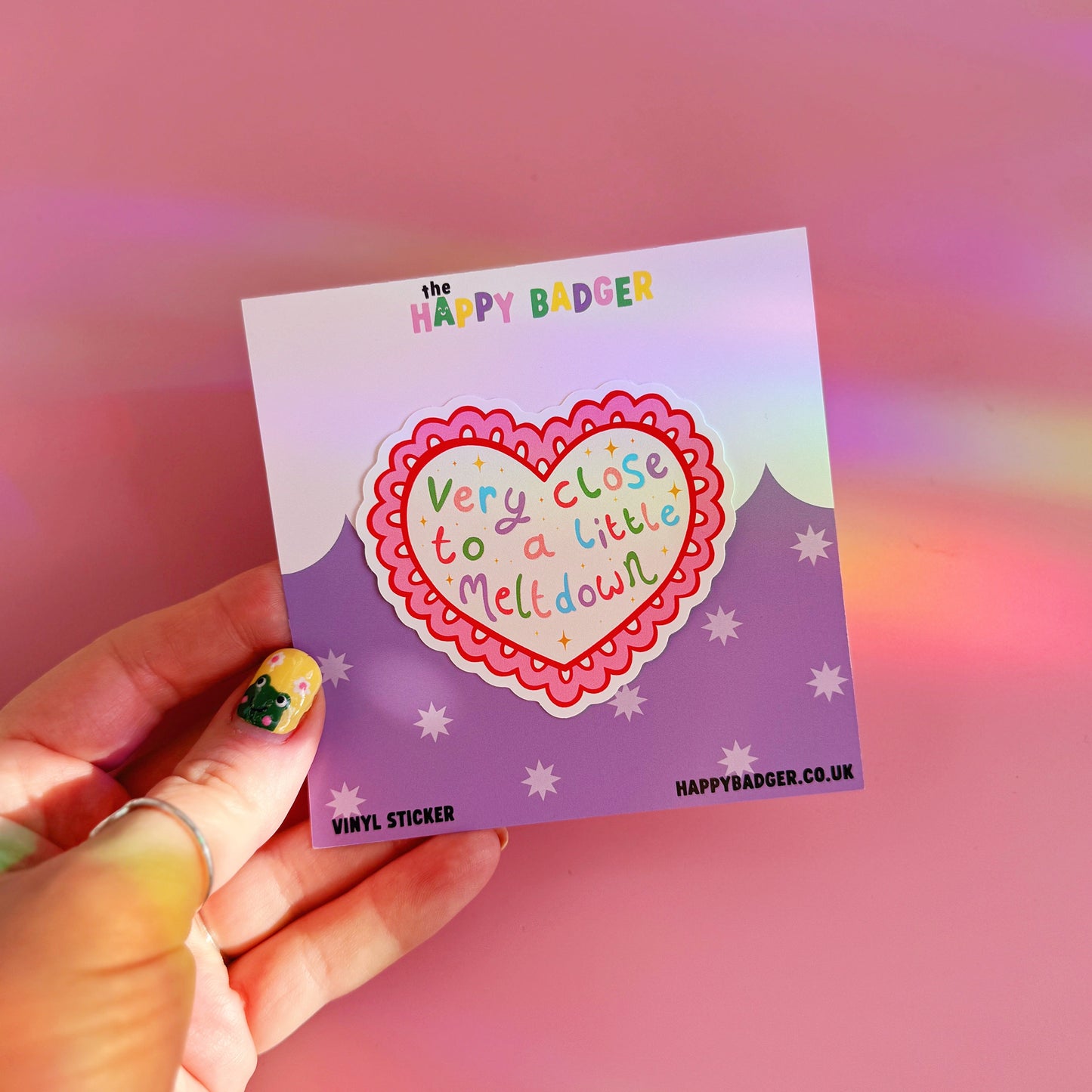 Heart-shaped sticker with a funny quote held by a hand against a pink background.