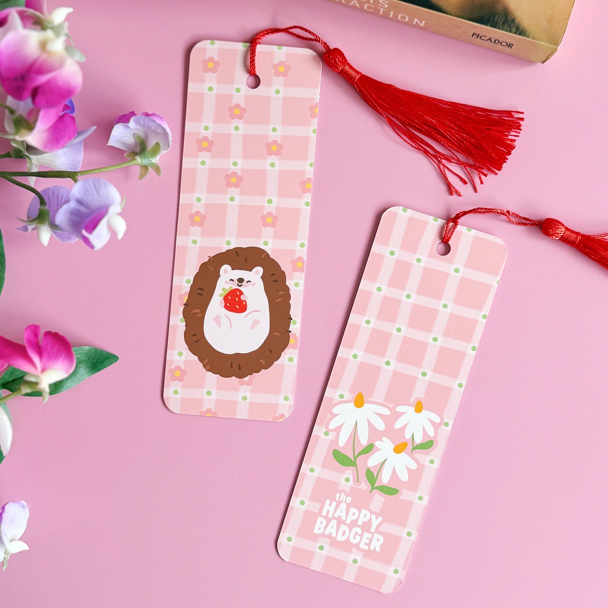 A pink bookmark with a red tassel, with little pink daisies dotted around a little hedgehog holding a strawberry. The back of the bookmark has the same pattern, but an illustration of daisies and the branding logo.