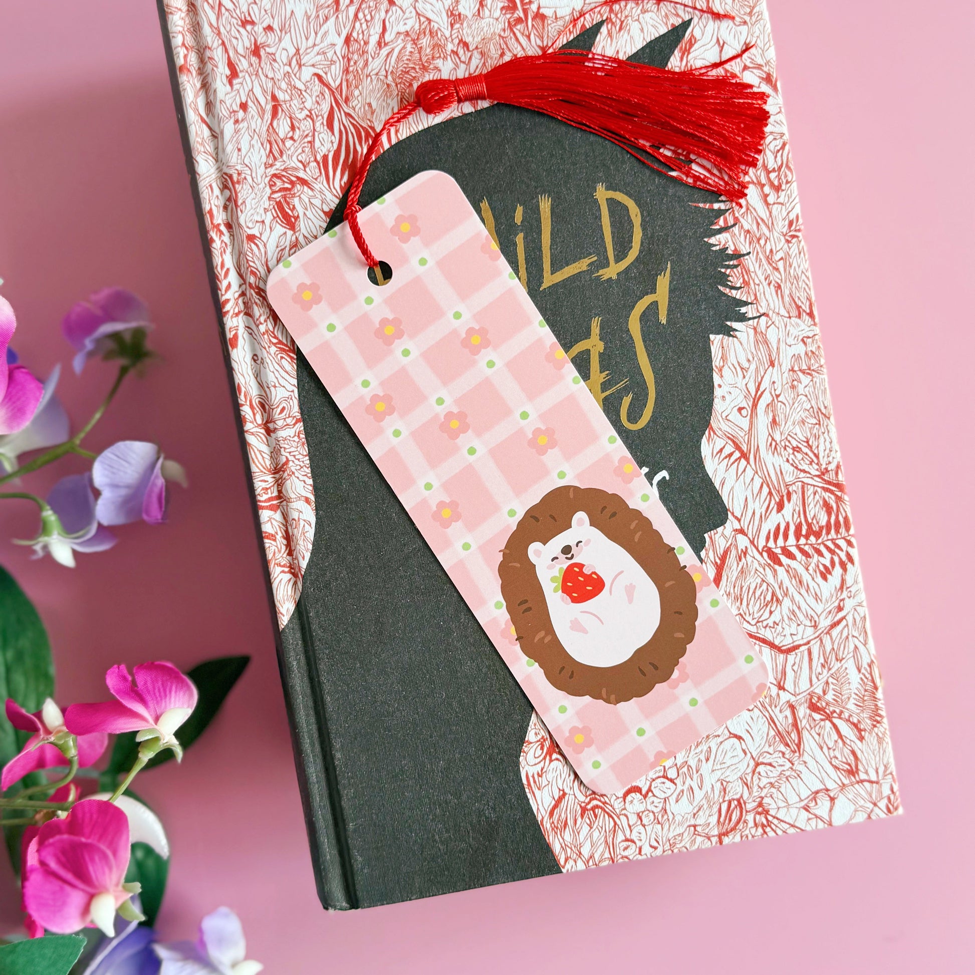 A pink bookmark with a red tassel, with little pink daisies dotted around a little hedgehog holding a strawberry. The back of the bookmark has the same pattern, but an illustration of daisies and the branding logo.