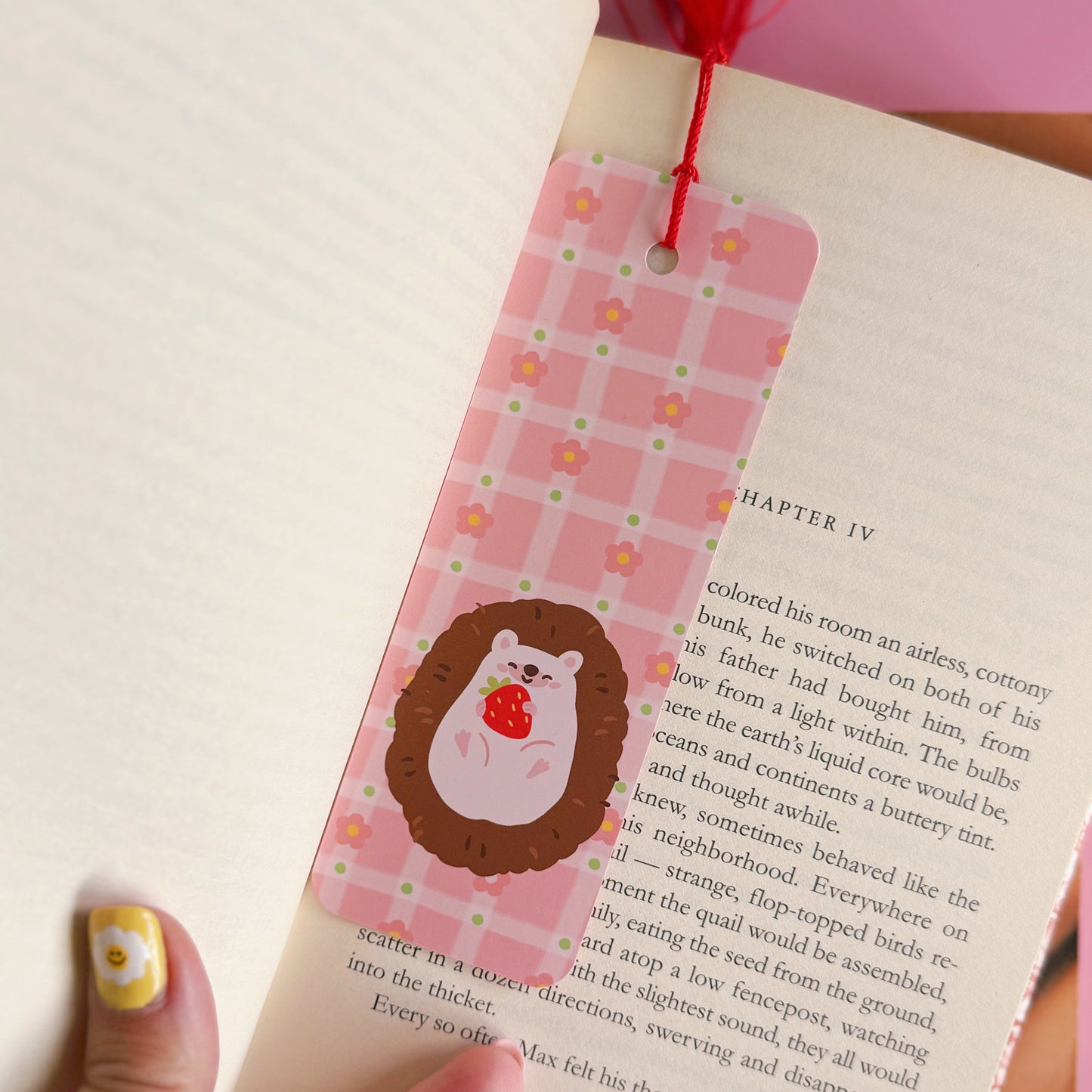 A pink bookmark with a red tassel, with little pink daisies dotted around a little hedgehog holding a strawberry. The back of the bookmark has the same pattern, but an illustration of daisies and the branding logo.