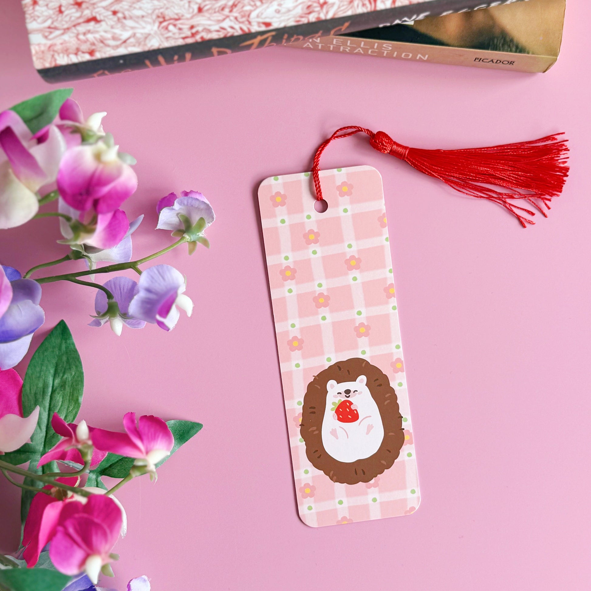 A pink bookmark with a red tassel, with little pink daisies dotted around a little hedgehog holding a strawberry. The back of the bookmark has the same pattern, but an illustration of daisies and the branding logo.