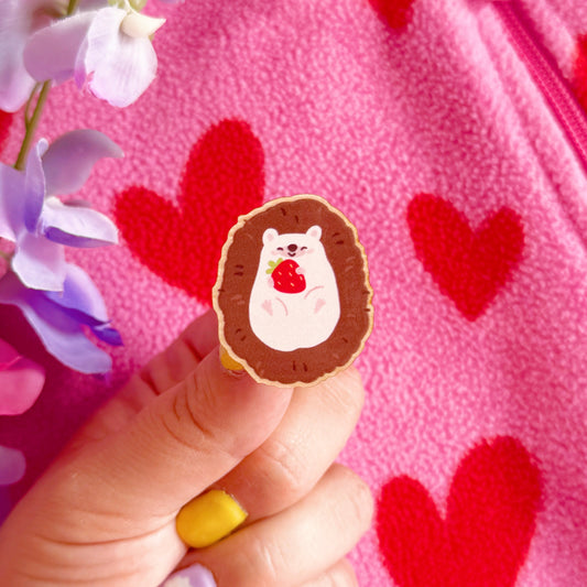 A wooden pin badge featuring a cute hedgehog curled up in a ball, with a little smiling face, holding a red strawberry in her arms.