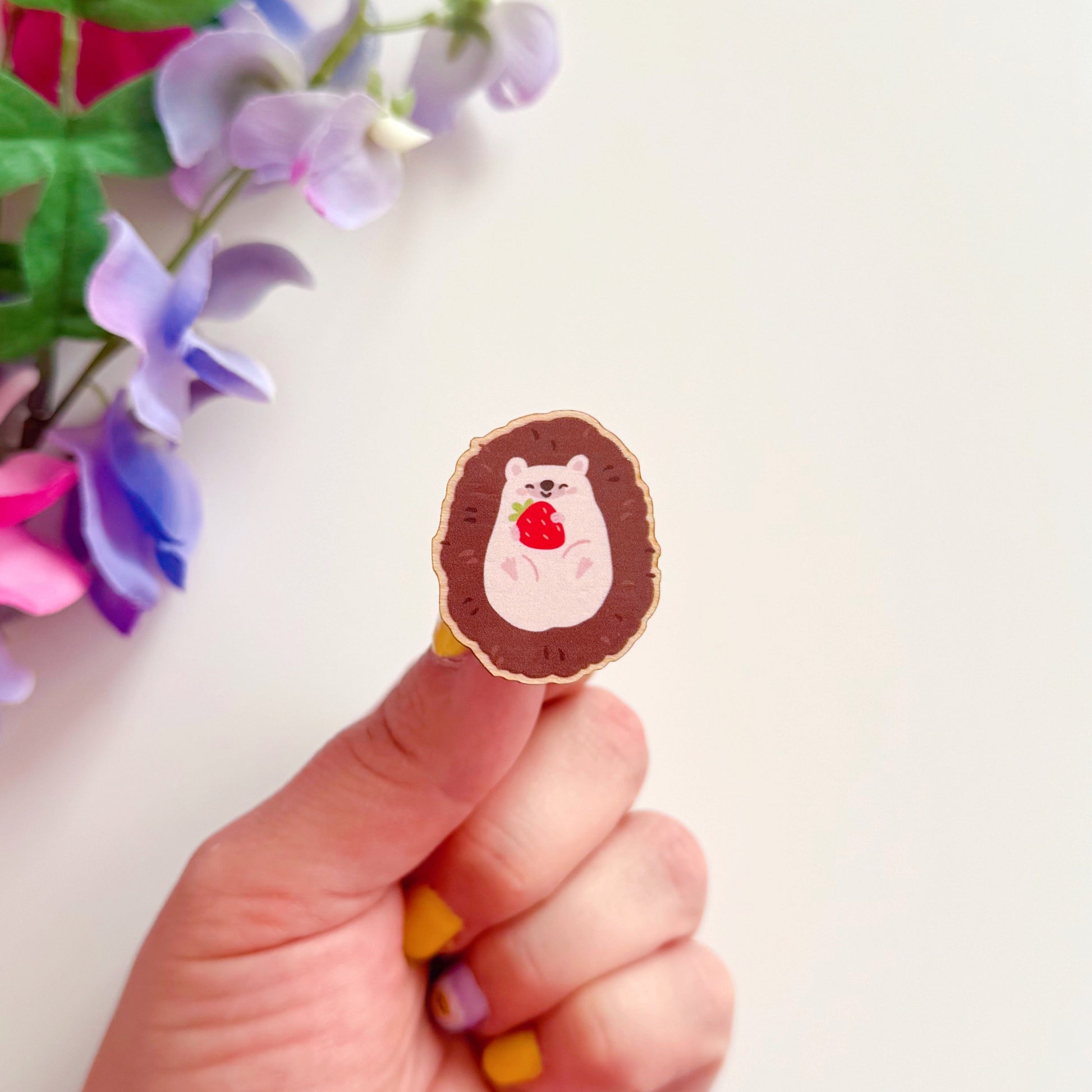 A wooden pin badge featuring a cute hedgehog curled up in a ball, with a little smiling face, holding a red strawberry in her arms.