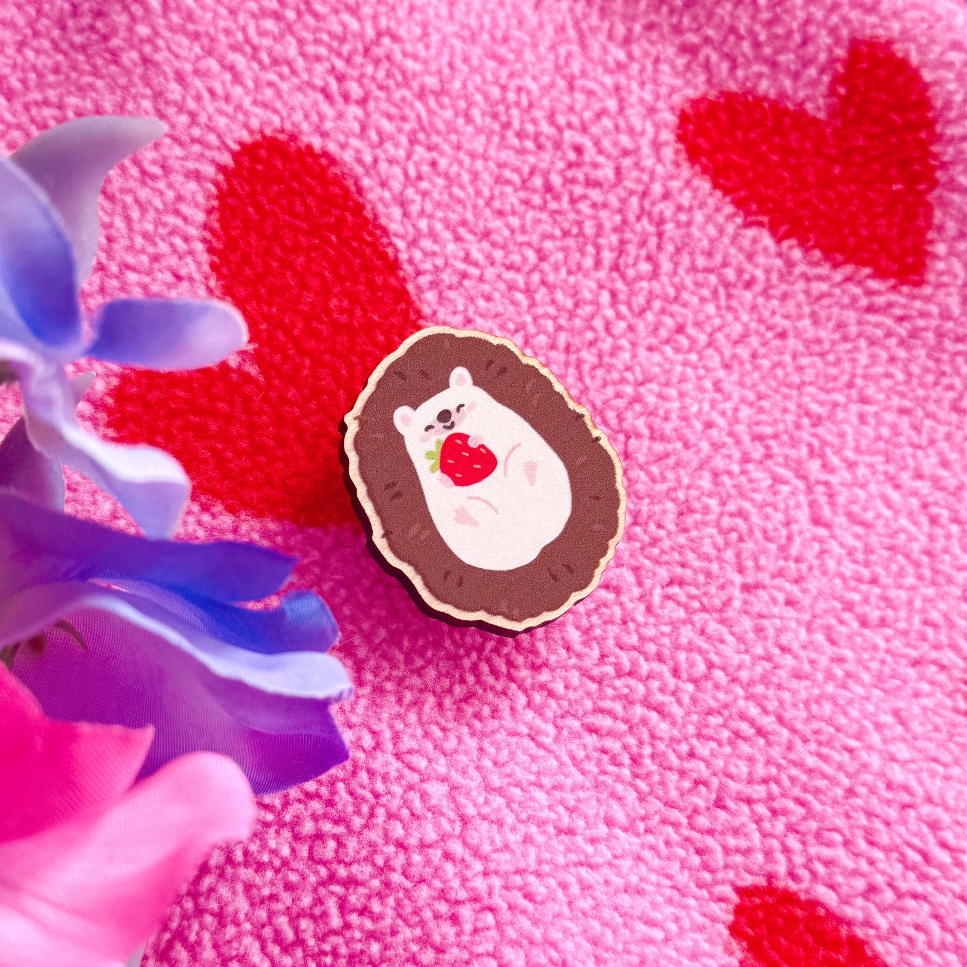 A wooden pin badge featuring a cute hedgehog curled up in a ball, with a little smiling face, holding a red strawberry in her arms.