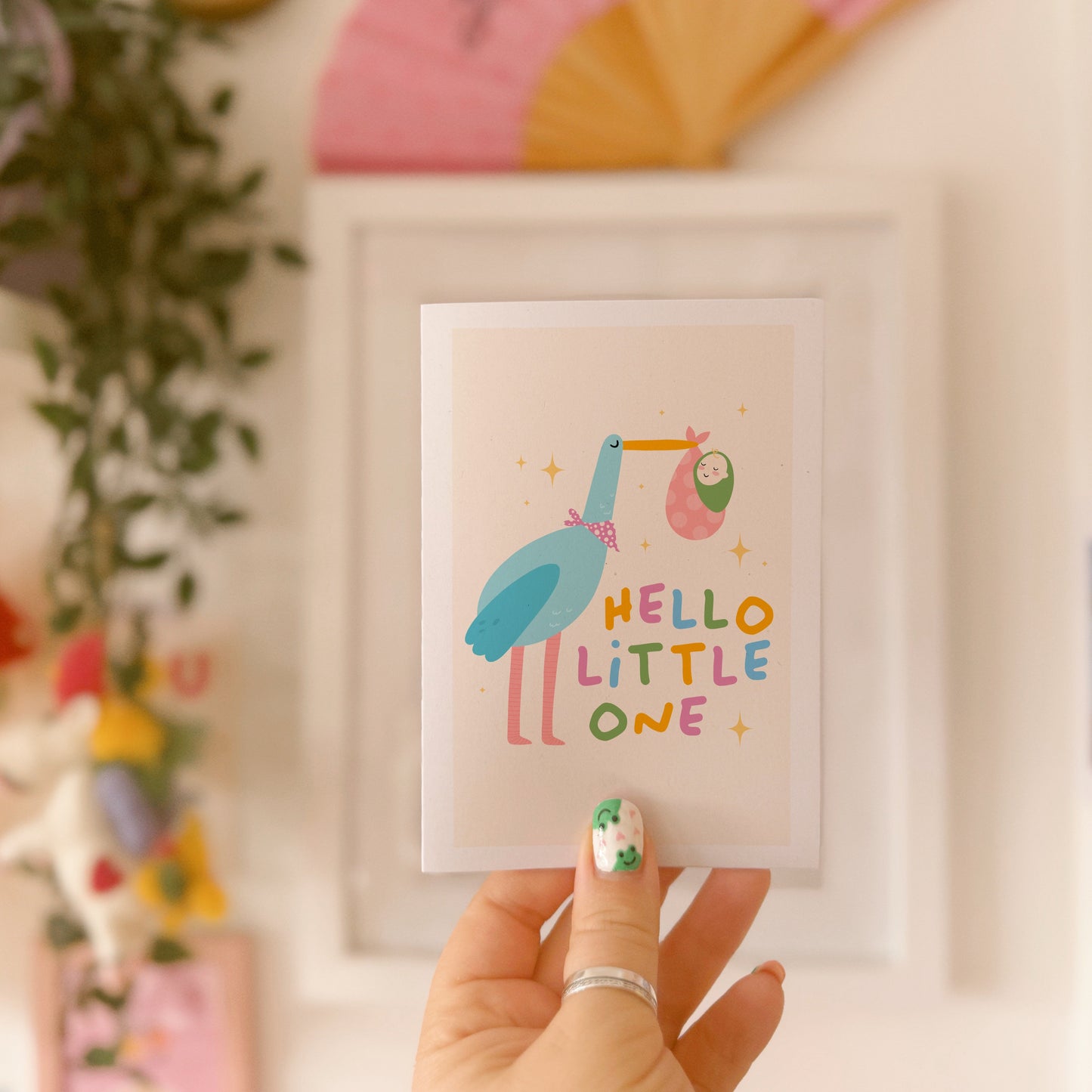 A greetings card with an illustration of a stalk carrying a baby, with the words 'hello little one' in colourful tones.