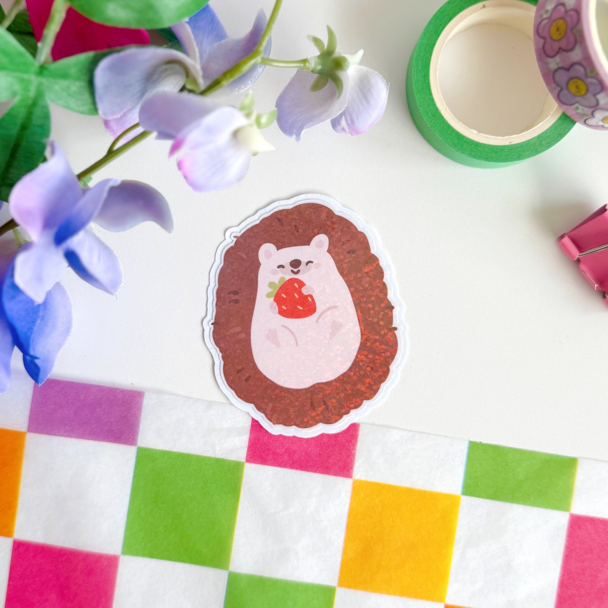 A glittery sticker of a small cute hedgehog holding a little strawberry in her hands.