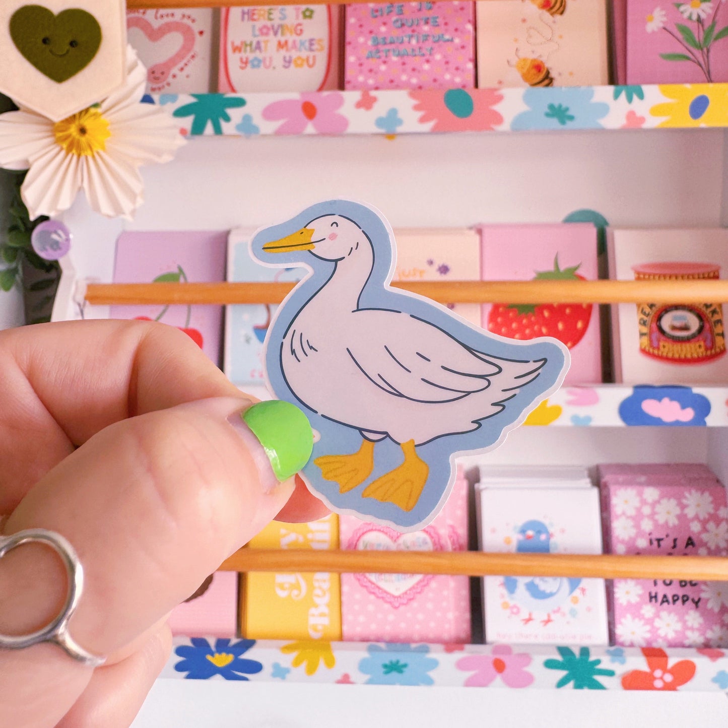 A sticker with an illustration of a white goose, smiling. The sticker has a blue border and the backing has an easy peel lip.
