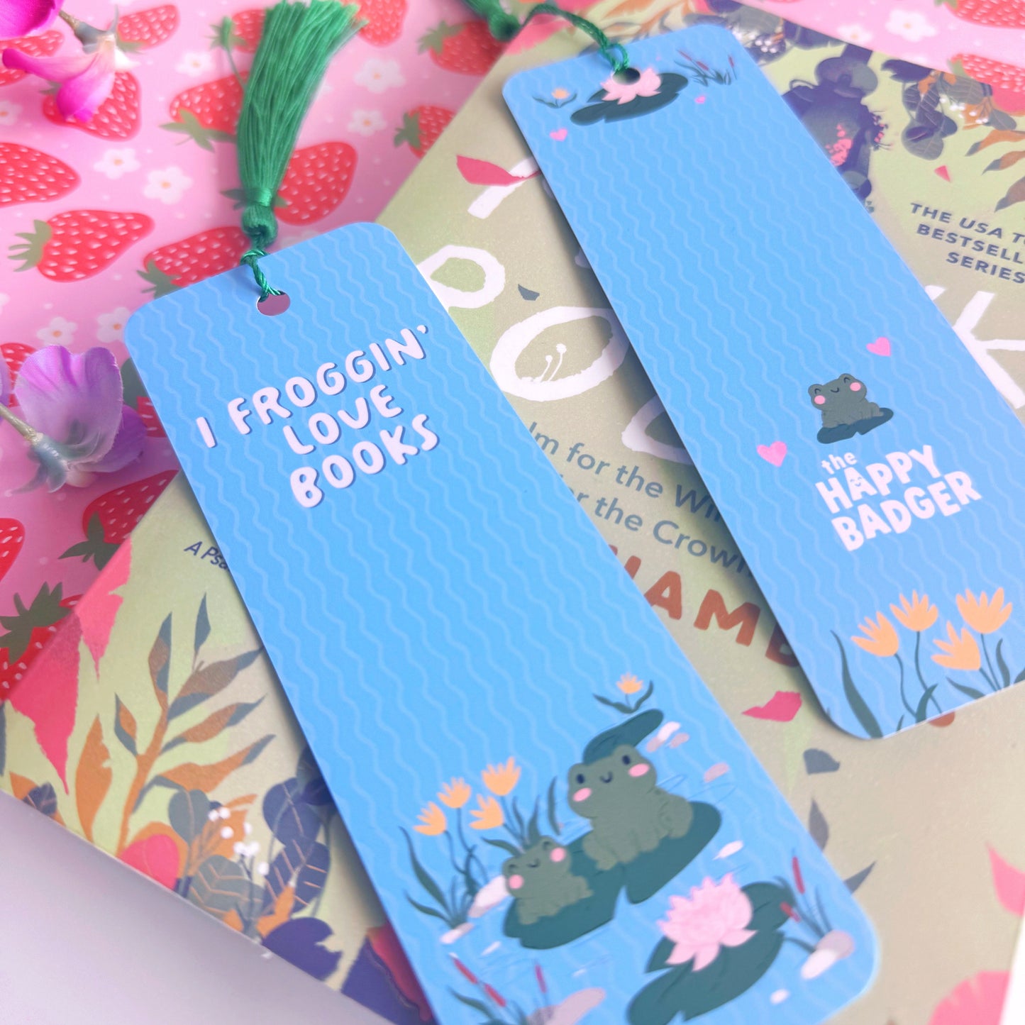 A blue bookmark with a cute frog illustration to both the front and back of the bookmark, on the front of the bookmark it says 'I Froggin' Love Books' and on the back of the bookmark is the brand logo.