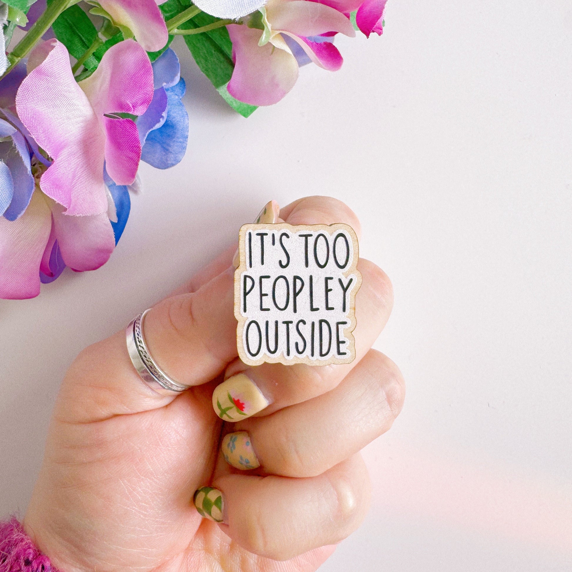 A wooden pin badge, that reads 'it's too peopley outside' on the front.