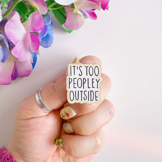 A wooden pin badge, that reads 'it's too peopley outside' on the front.