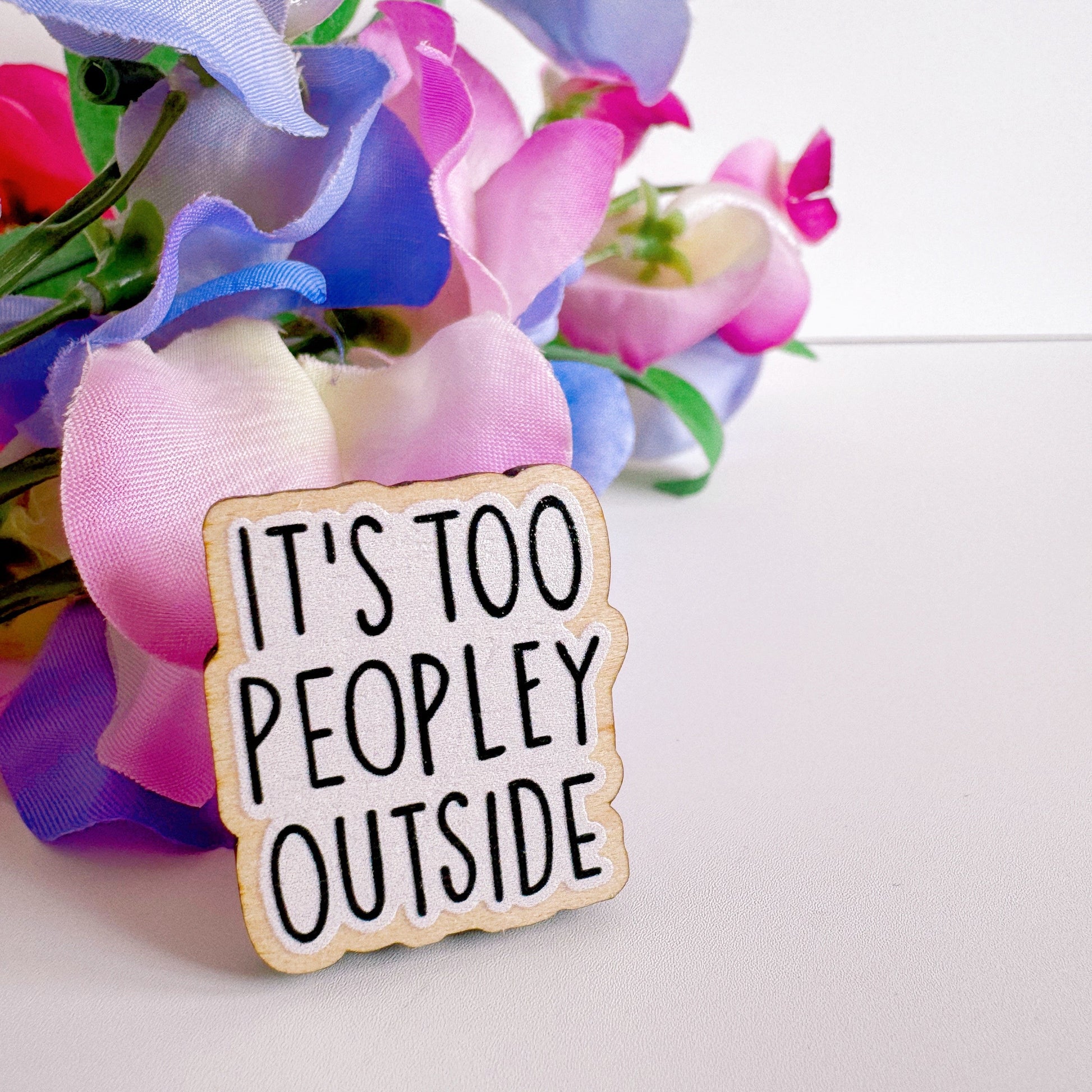 A wooden pin badge, that reads 'it's too peopley outside' on the front.