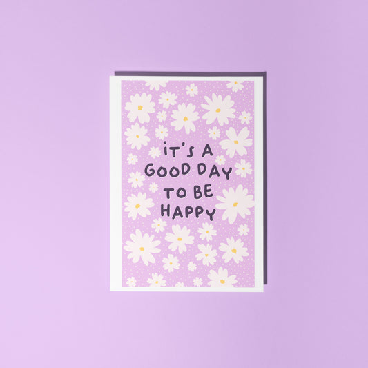 A purple card, with a white daisy pattern all over and the words 'it's a good day to be happy' in the centre.