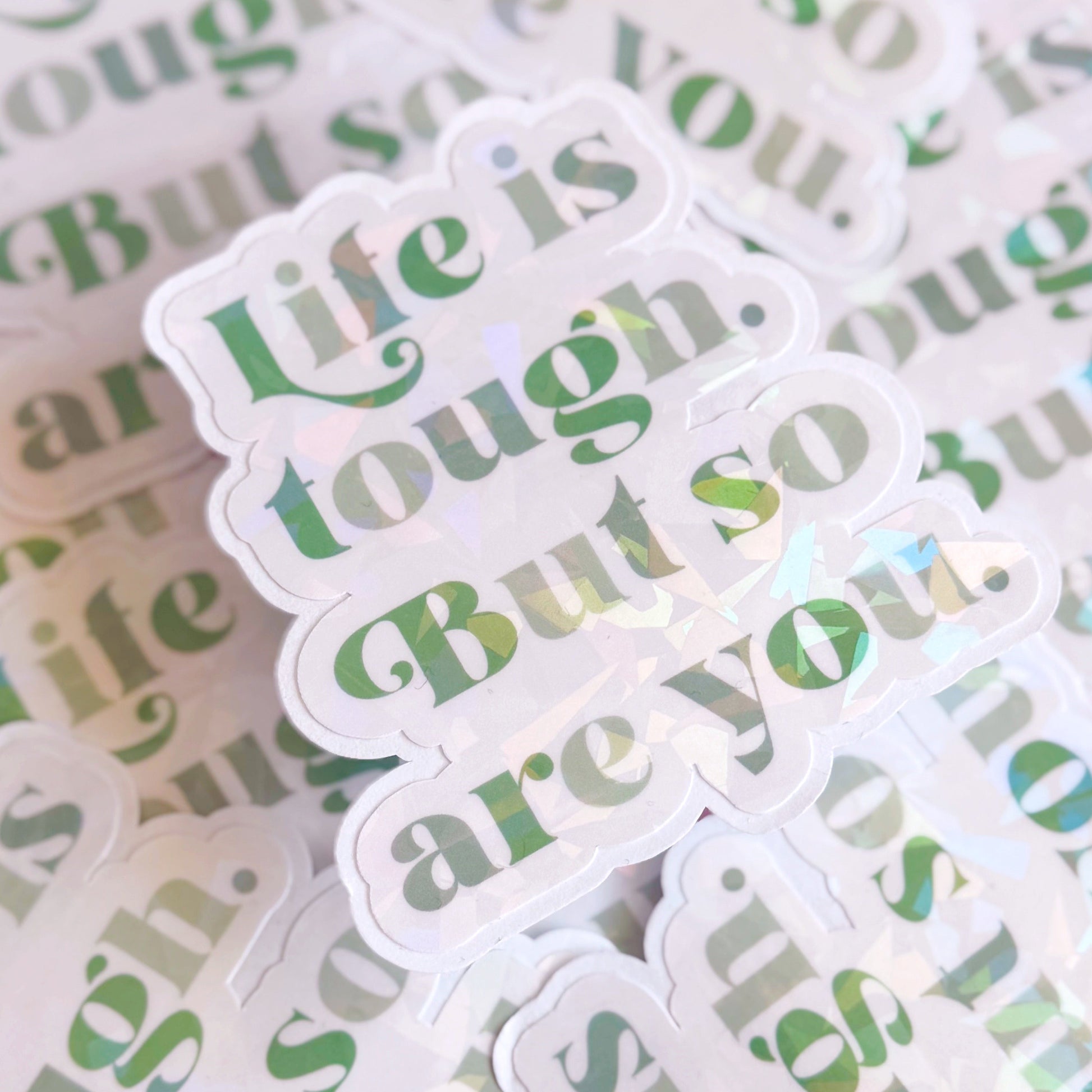A green holographic finish sticker, with green text saying 'life is tough but so are you'.
