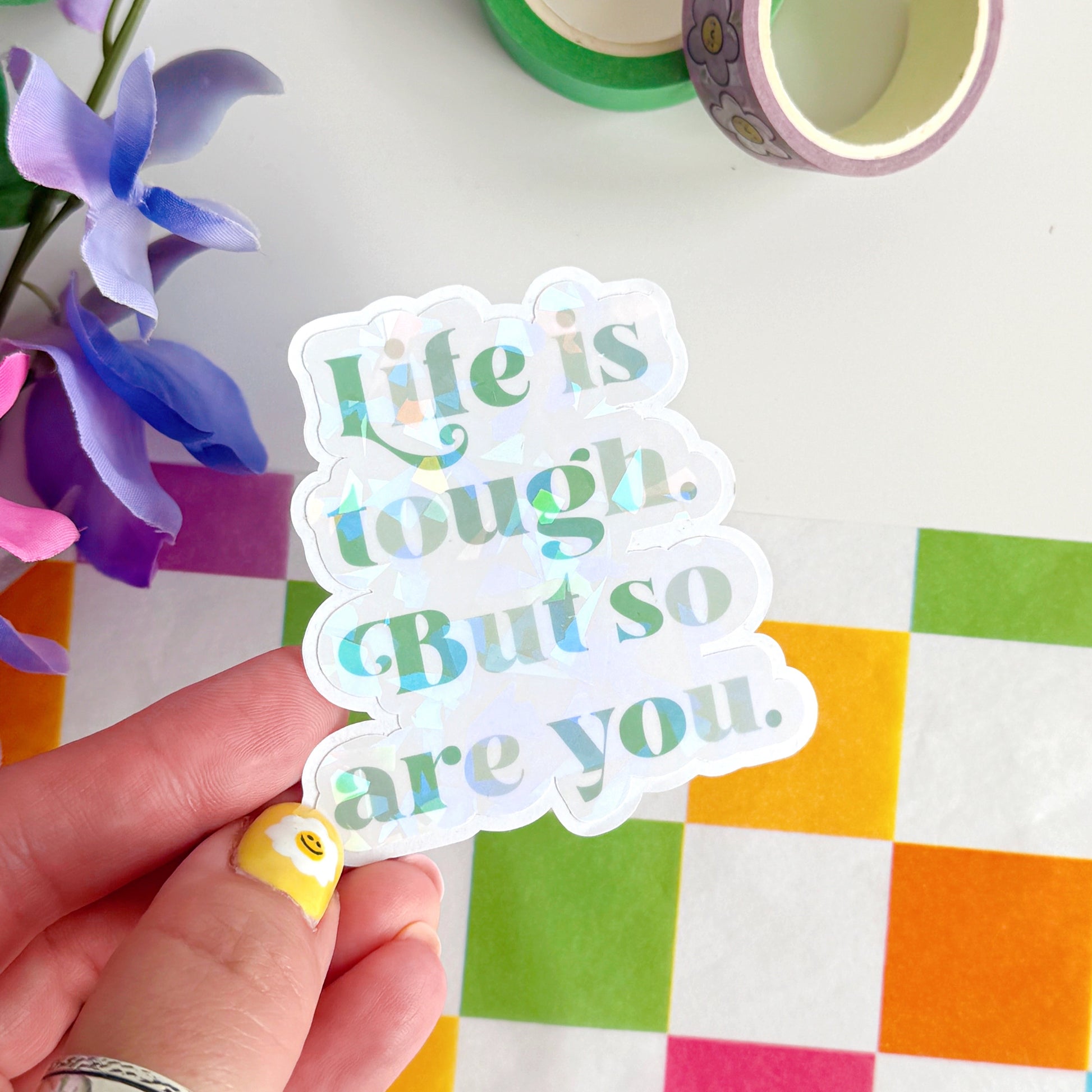 A green holographic finish sticker, with green text saying 'life is tough but so are you'.