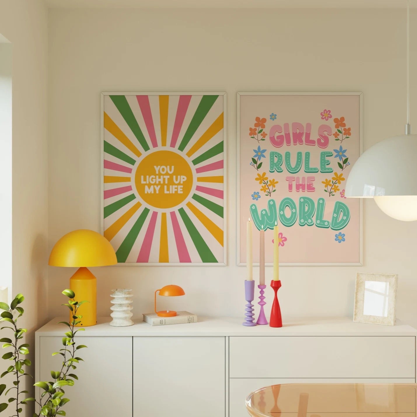 A bright vibrant print saying 'Girls Rule The World' in pink and teal, with flowers illustrated around the sides. Shown next to a colourful pink yellow and green print saying 'You Light Up My Life.'