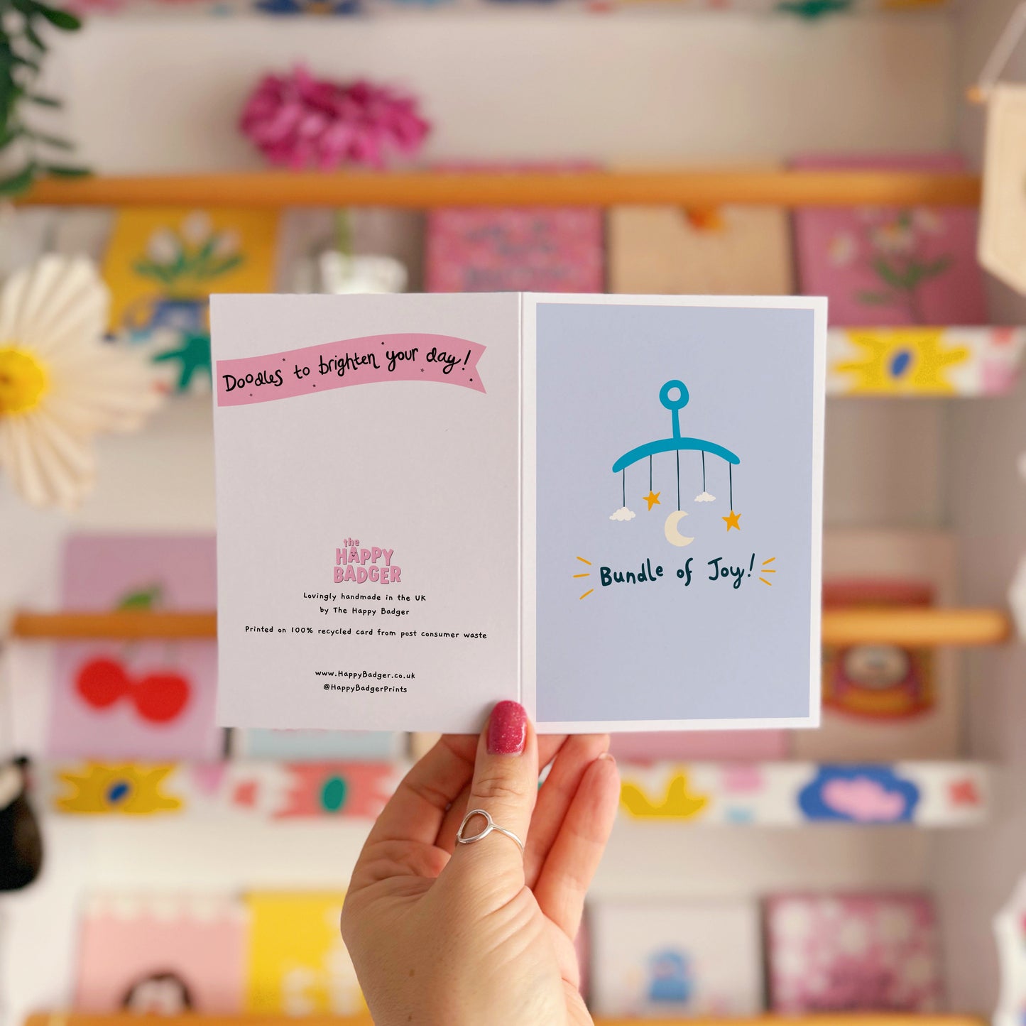 A blue greeting card with an illustration of a mobile. Underneath the illustration is hand written text that reads 'bundle of joy!'.