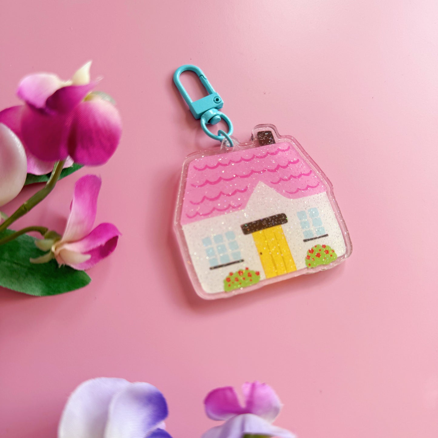 House-shaped keychain with a pink roof and yellow door on a pink background with flowers.