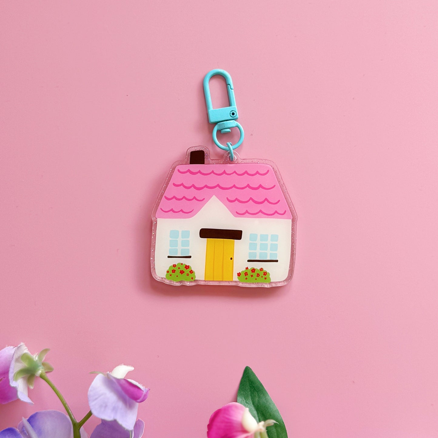 House-shaped keychain with a blue clip on a pink background with flowers at the bottom.