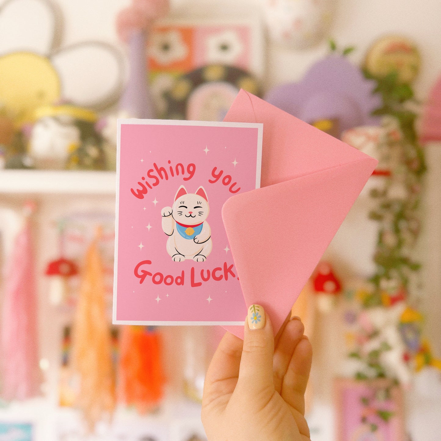 A pink greeting card with a lucky cat illustration and text that reads 'Wishing you Good Luck!'.