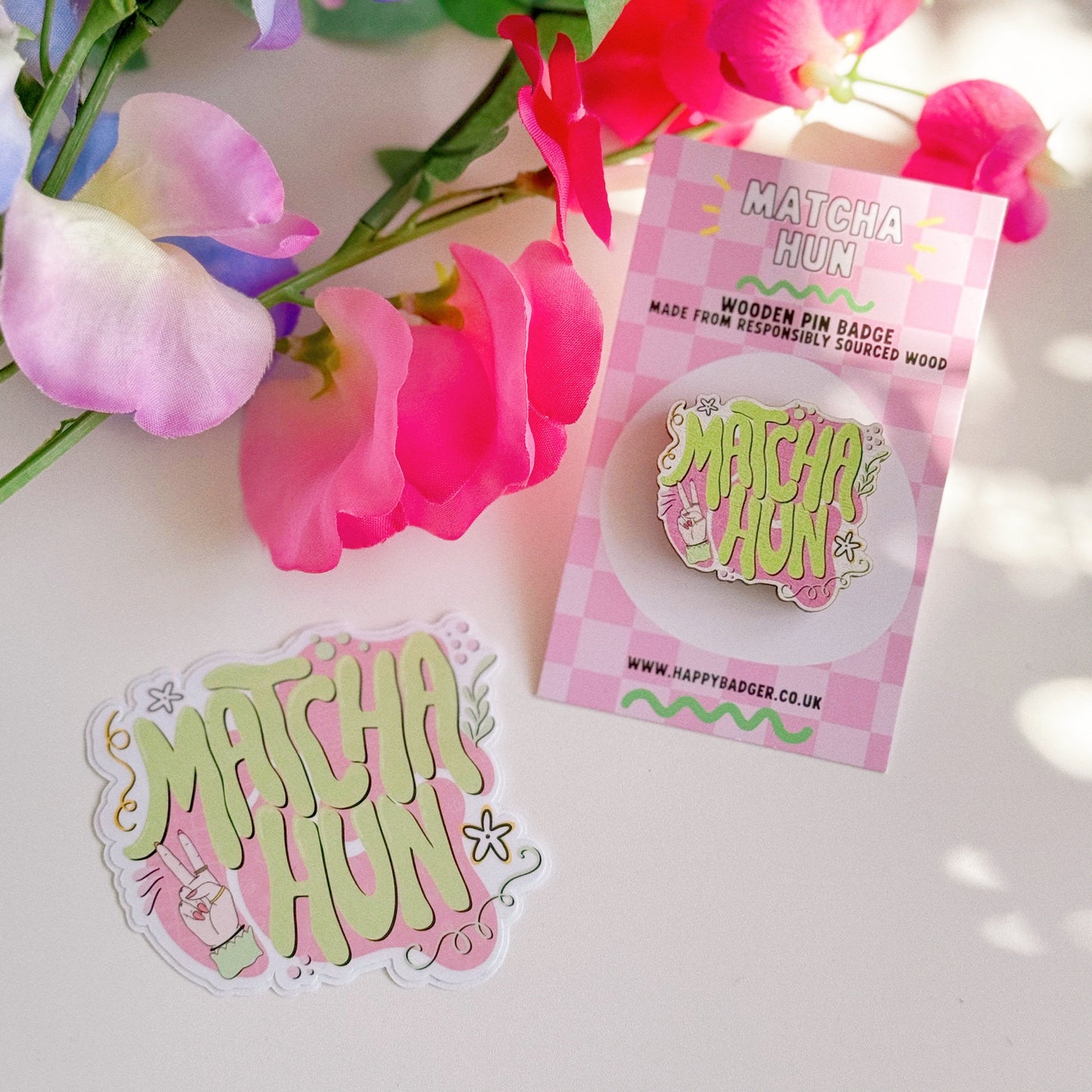 An eco friendly wooden pin badge, with a pink and green design that says 'matcha hun'. Shown next to a glittery holographic sticker of the same design.