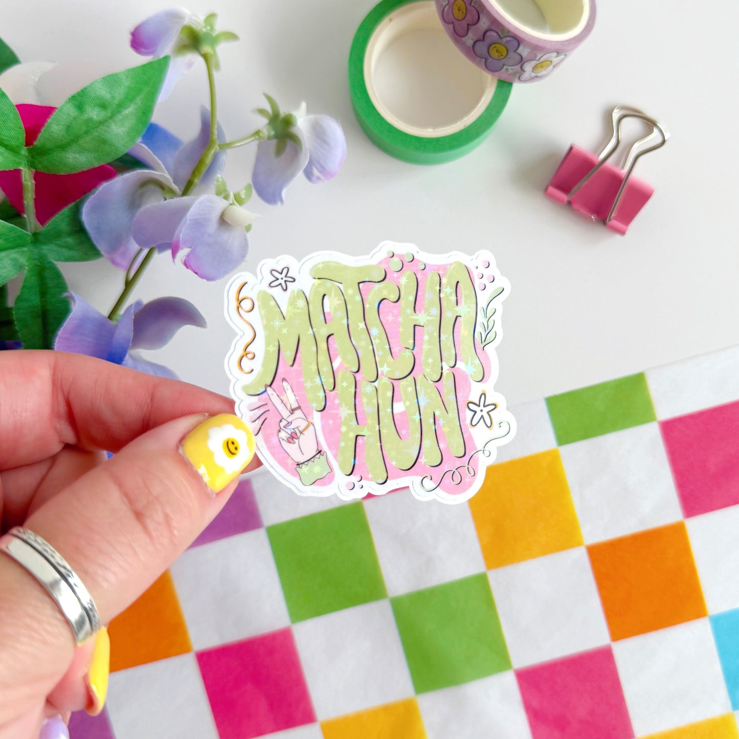 A holographic sticker with text saying 'Matcha Hun' in funky bubble writing and other hand illustrated elements around the text. The colours are pink and green.