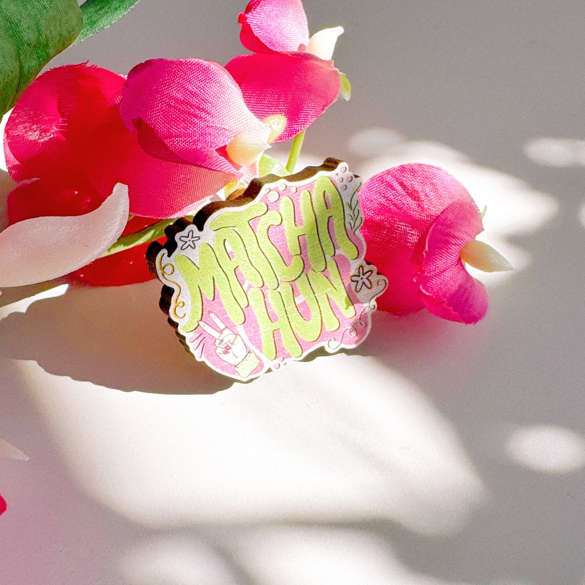 An eco friendly wooden pin badge, with a pink and green design that says 'matcha hun'.