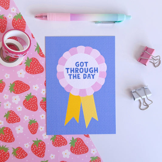 A blue postcard, with a rosette in pink and yellow, saying got through the day.