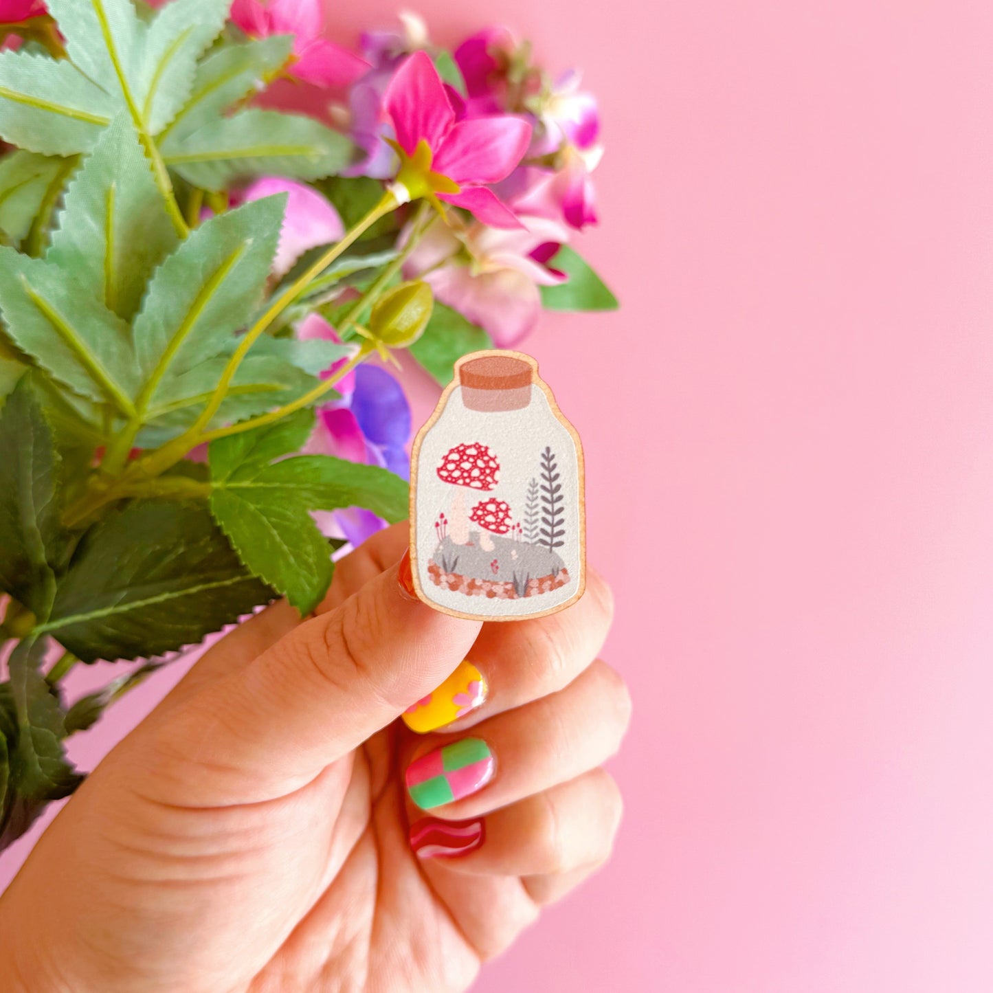 A terrarium jar illustration, with a mini woodland scene with mushrooms and ferns inside, on an eco friendly wooden pin badge.