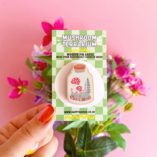 A terrarium jar illustration, with a mini woodland scene with mushrooms and ferns inside, on an eco friendly wooden pin badge. Shown on a jazzy bright green pin backing card.