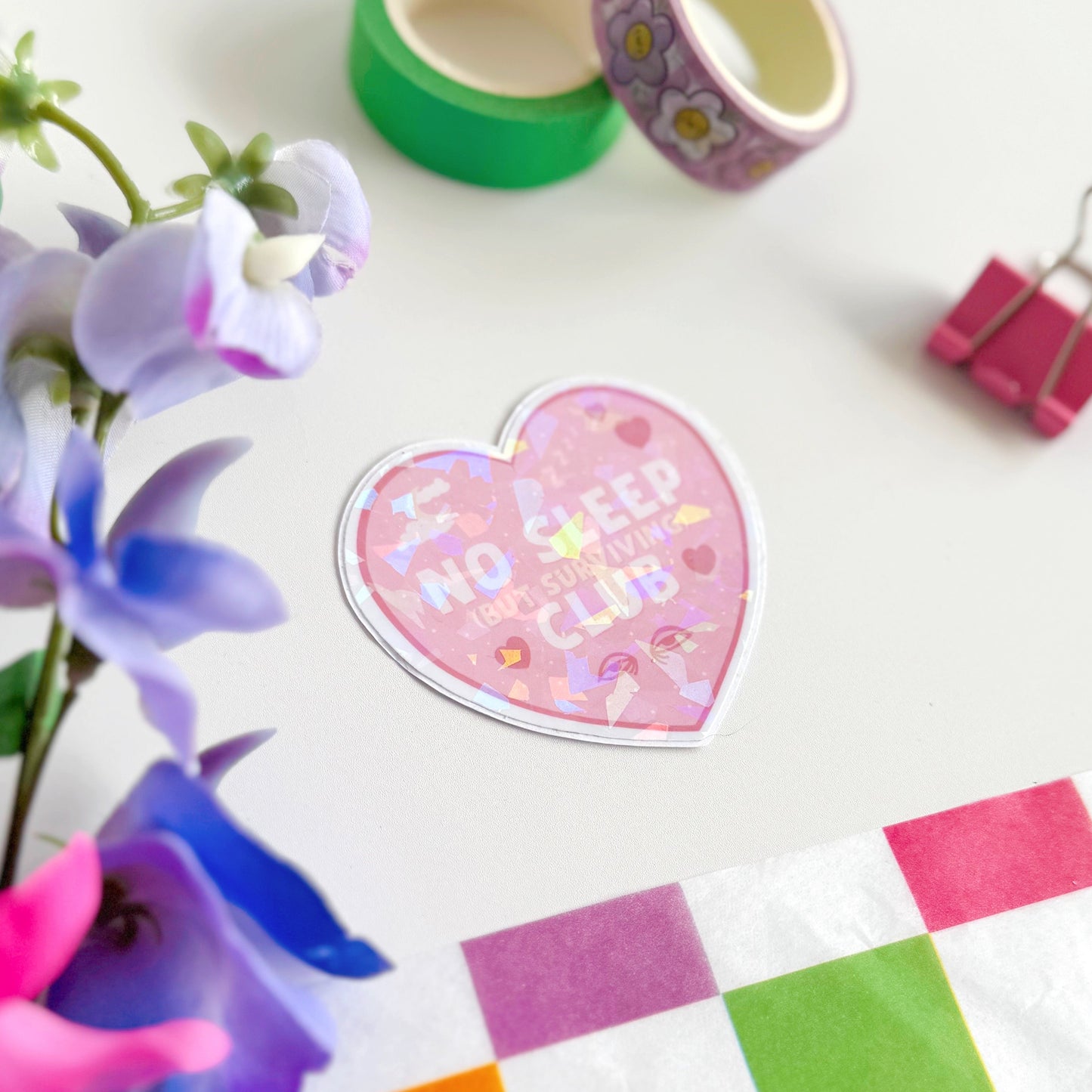 A pink holographic sticker, in the shape of a heart, with the words 'No sleep (but surviving) club' in the centre. Around the text are little doodles, like tired eyes, hearts and a moon behind clouds.