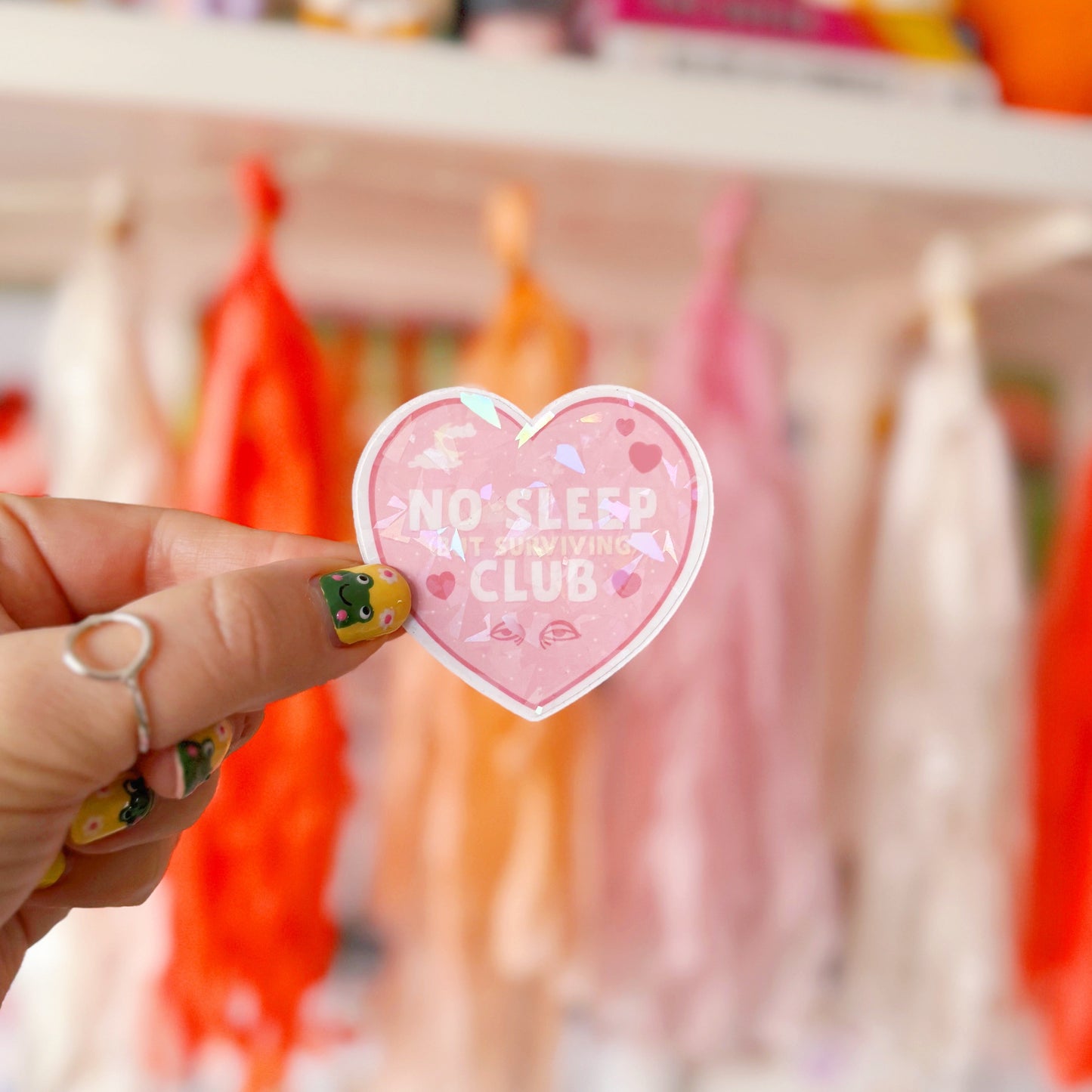 A pink holographic sticker, in the shape of a heart, with the words 'No sleep (but surviving) club' in the centre. Around the text are little doodles, like tired eyes, hearts and a moon behind clouds.